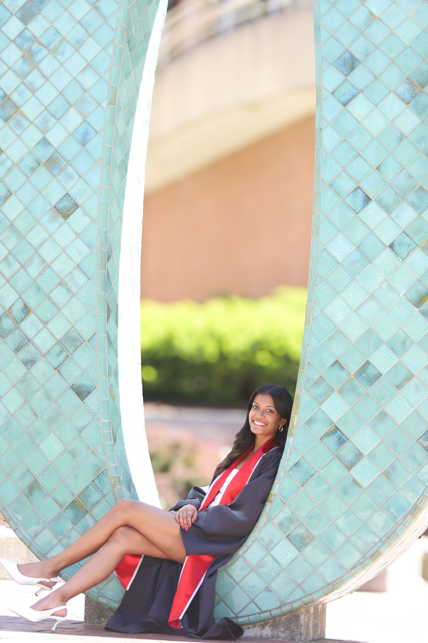 A person sits in a curved architectural opening with turquoise mosaic tiles, wearing graduation attire on a sunny day.