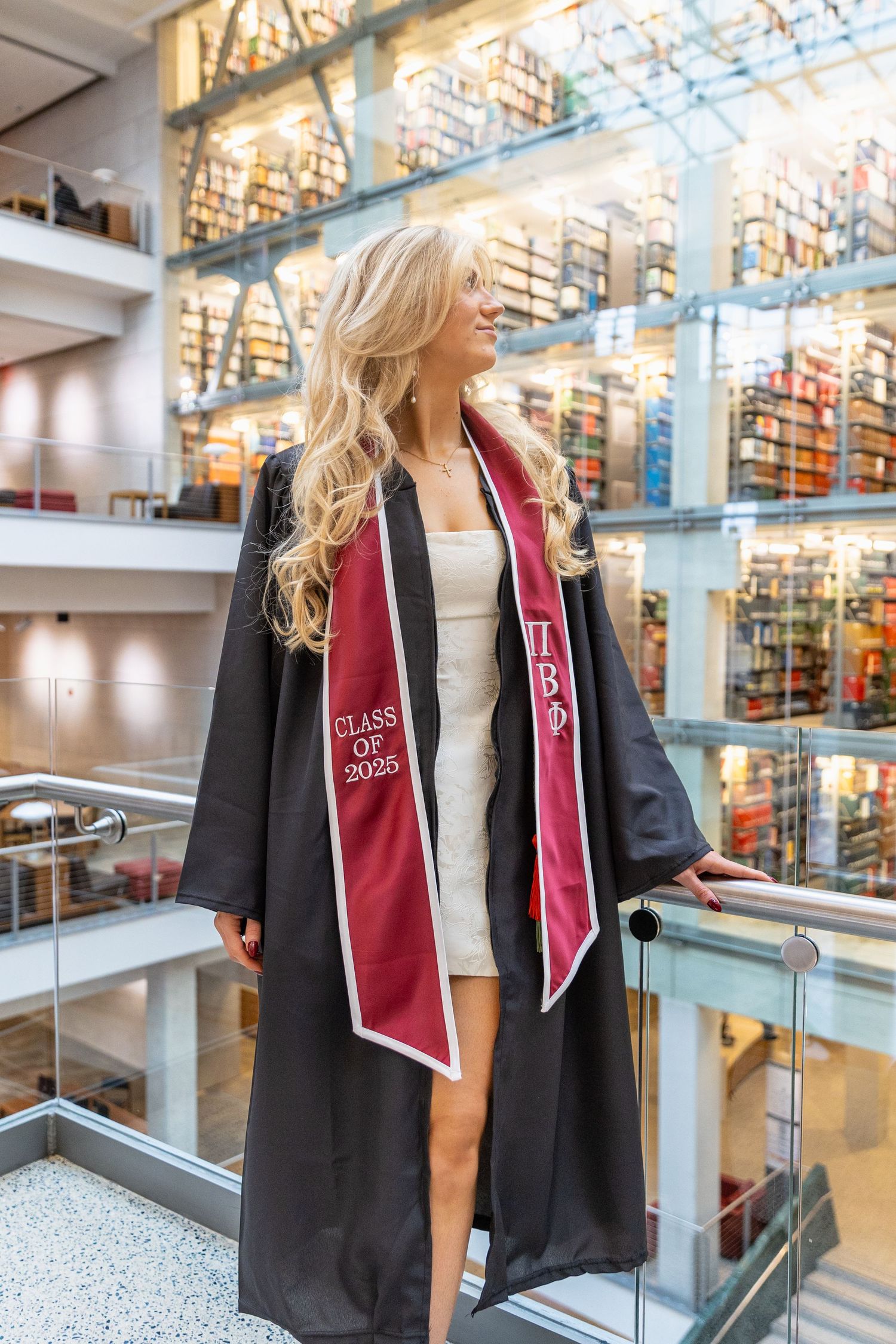 Graduate wearing black cap and gown with red stole poses in modern library atrium with glass walls and bookshelves.