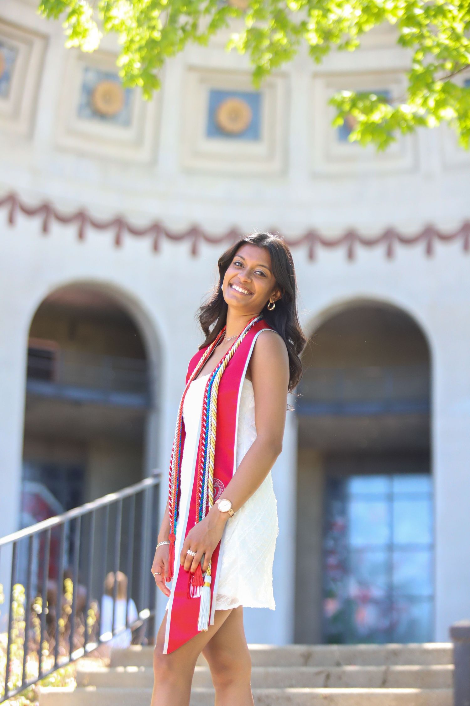 Graduate in white dress and red graduation sash smiles while standing on steps of building with arched columns.