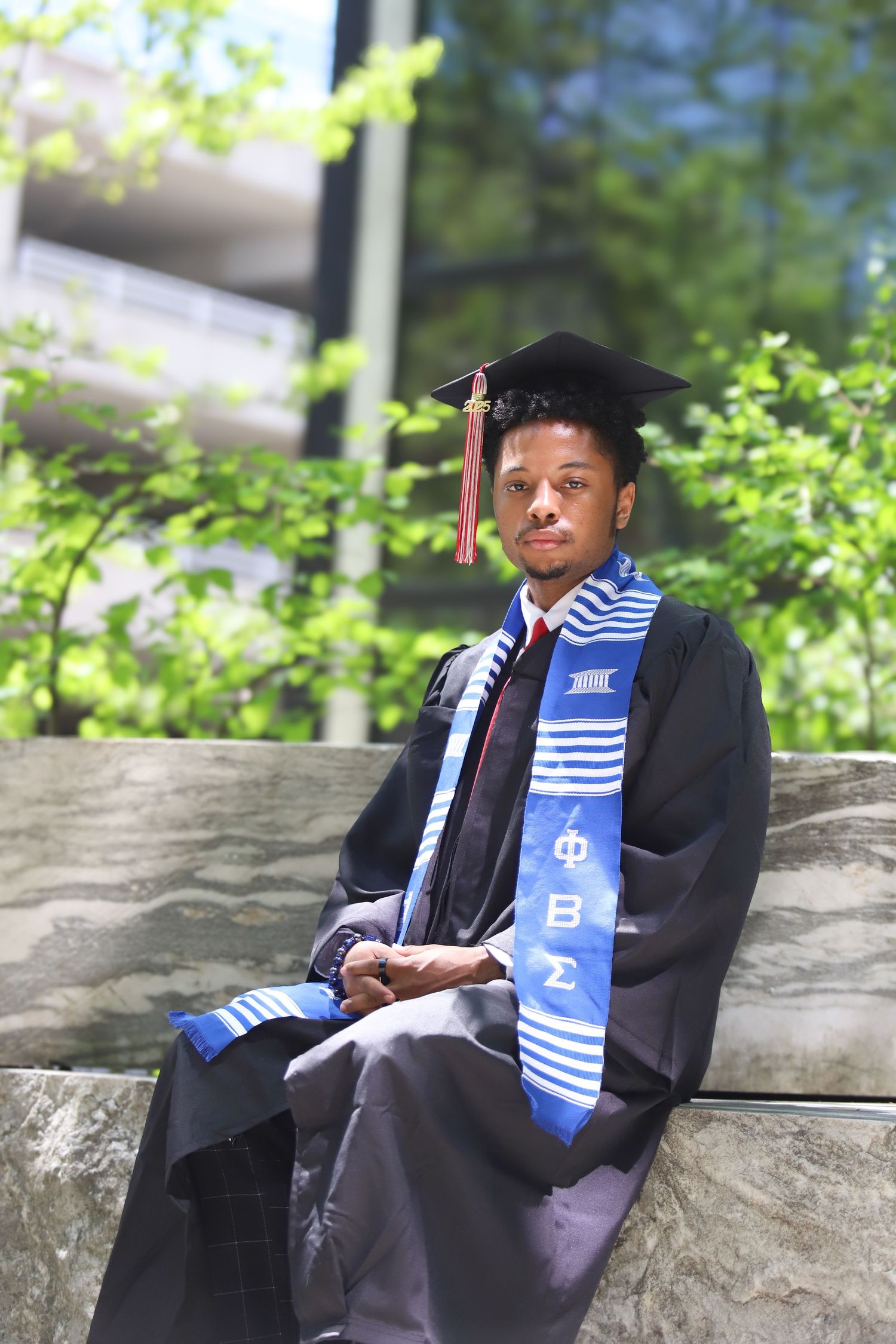 Graduate in cap and gown with blue and white stole sits on stone wall during outdoor graduation photo session.