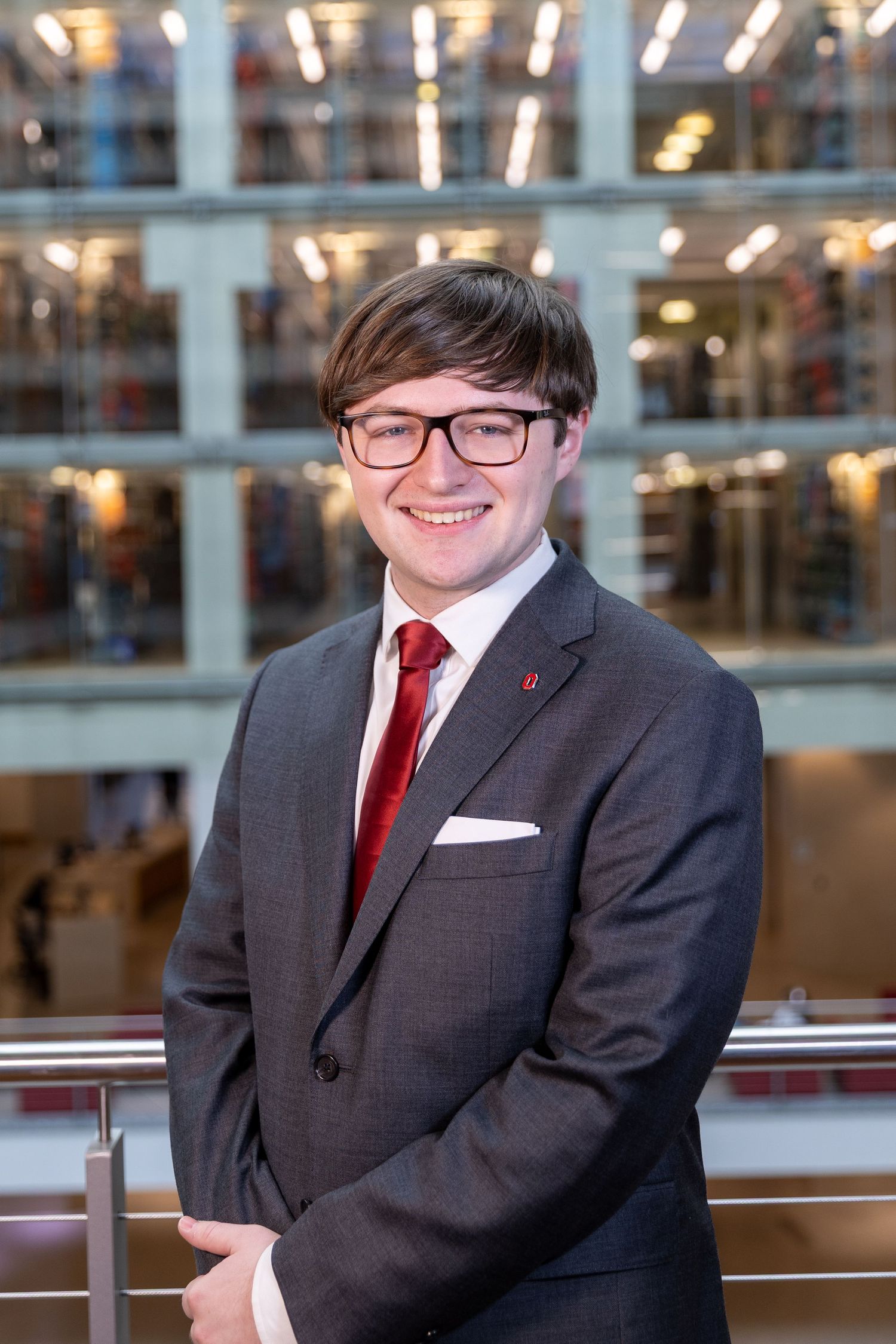 Professional portrait of someone in a dark gray suit with red tie and glasses standing in front of a modern office building interior.