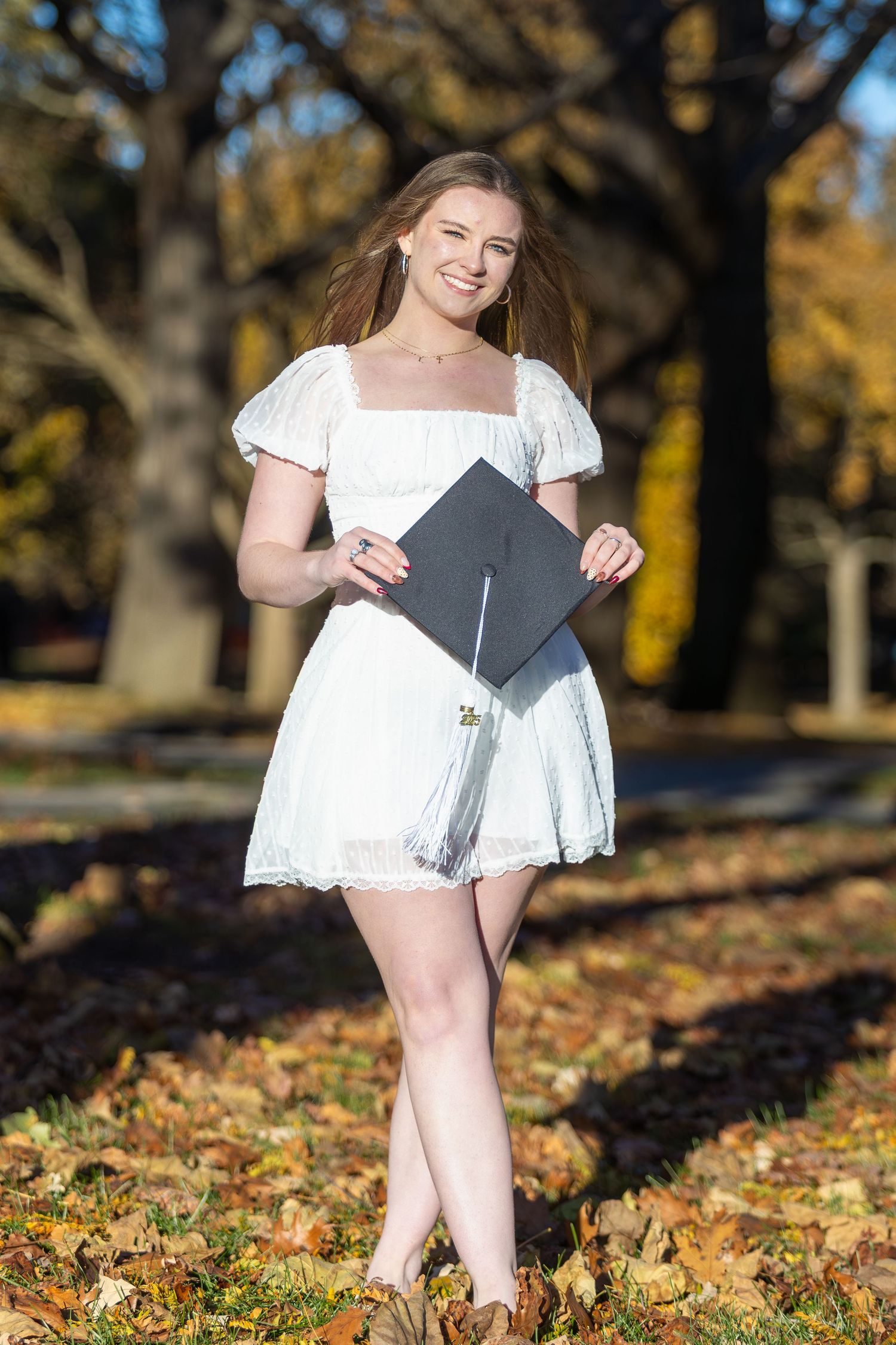 Graduate in white dress sits among autumn leaves for graduation photos.