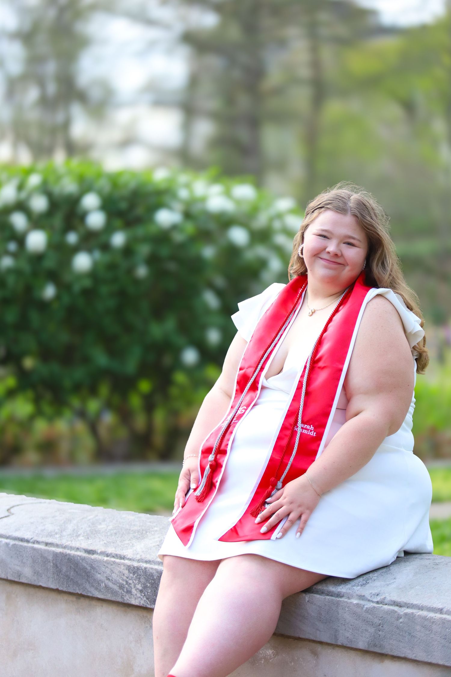 A figure in a red graduation stole reclines on a stone ledge outdoors with greenery in the background.