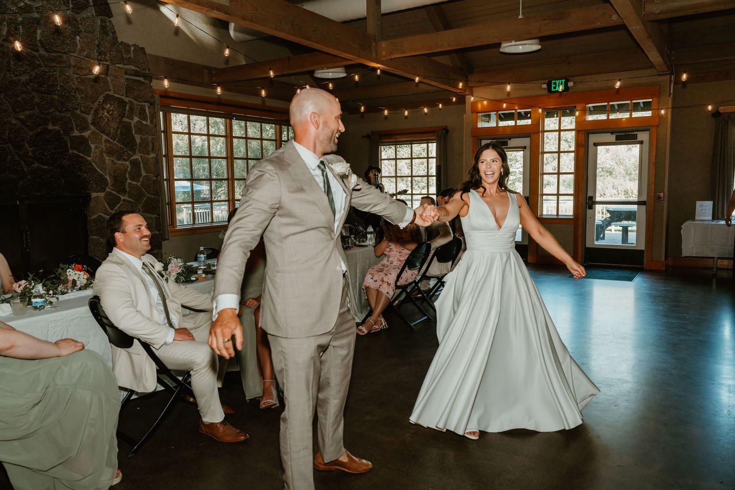 Bride and groom share first dance in rustic wedding venue with string lights and stone fireplace.
