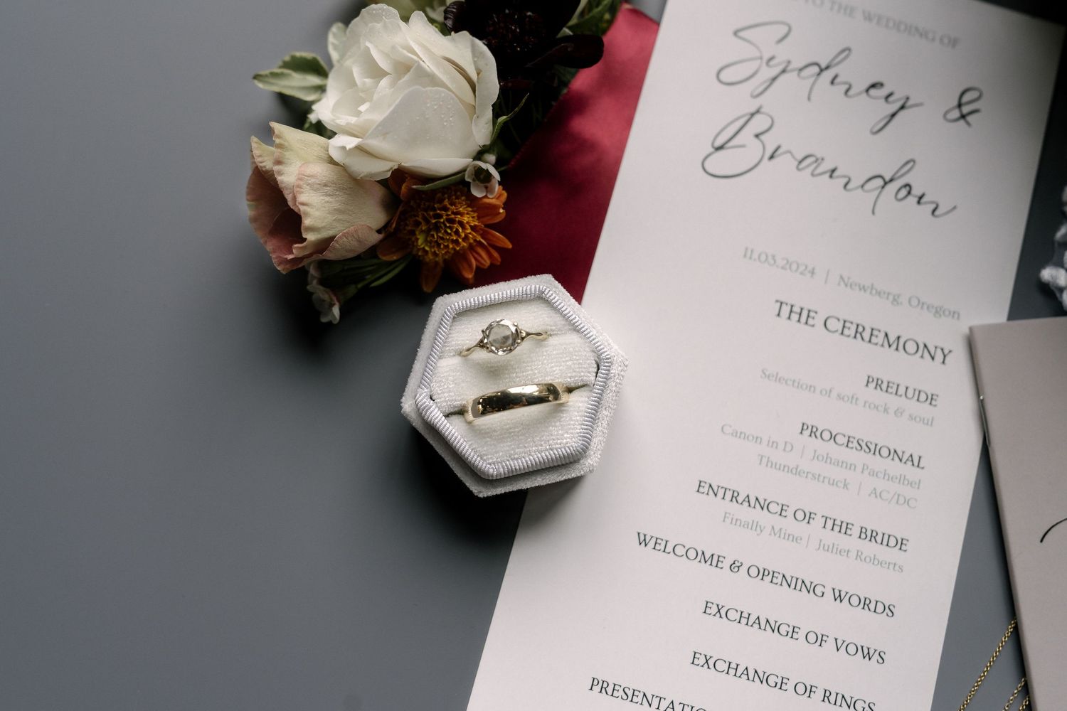 Wedding rings in a velvet box beside a ceremony program for Sydney and Brandon, with white and red floral bouquet on gray background.