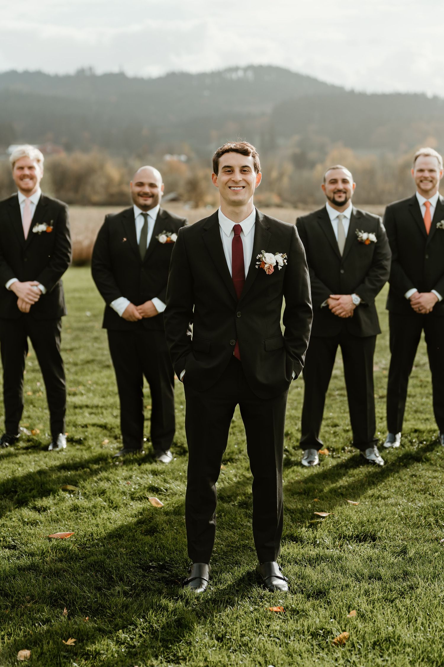 Groom in black suit with burgundy tie stands before groomsmen in matching suits on green lawn with mountain backdrop.