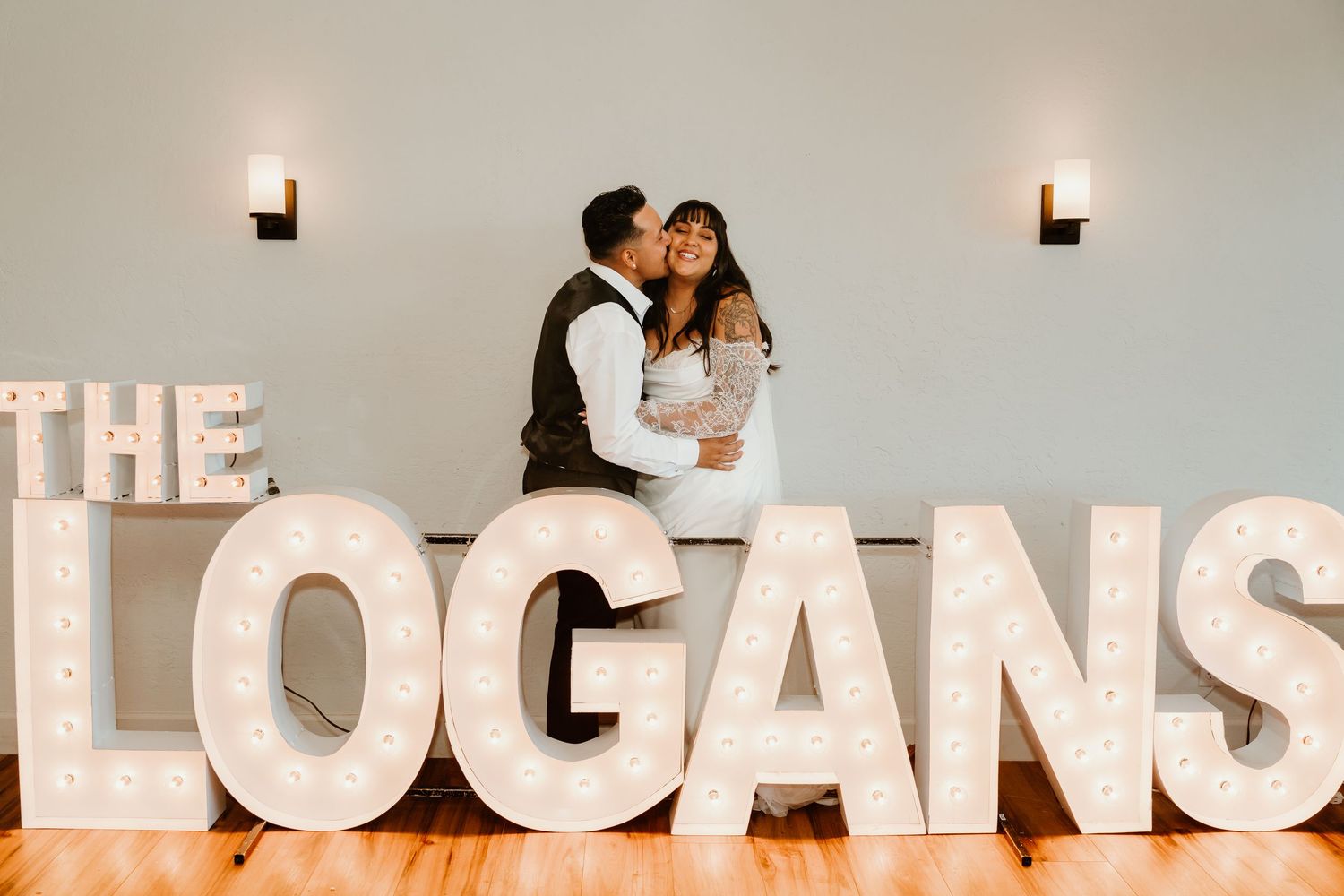 Newlyweds share a kiss behind large marquee light-up letters spelling THE LOGANS at their wedding reception.