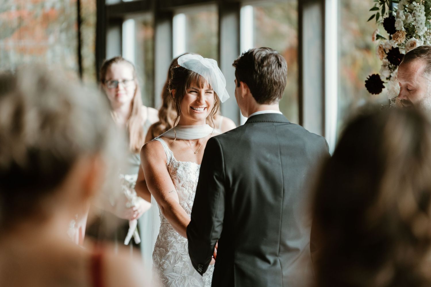 Smiling bride in lace gown and veil exchanges vows with groom during indoor wedding ceremony.