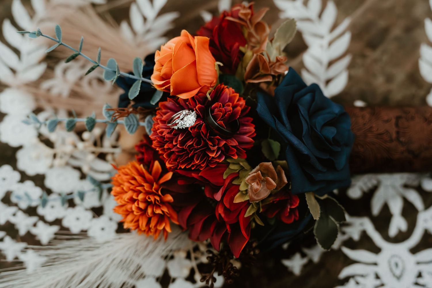 Vibrant fall wedding bouquet with orange dahlias, deep red mums, and navy roses on lace fabric.