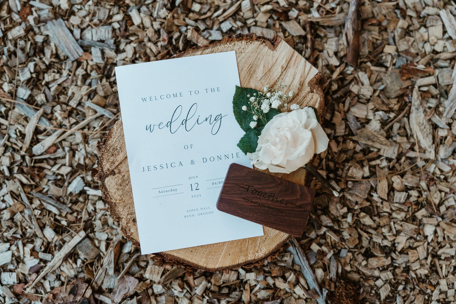 Wedding program for Jessica and Ronnie displayed on wood slice with white flowers and wooden ring box on mulch.