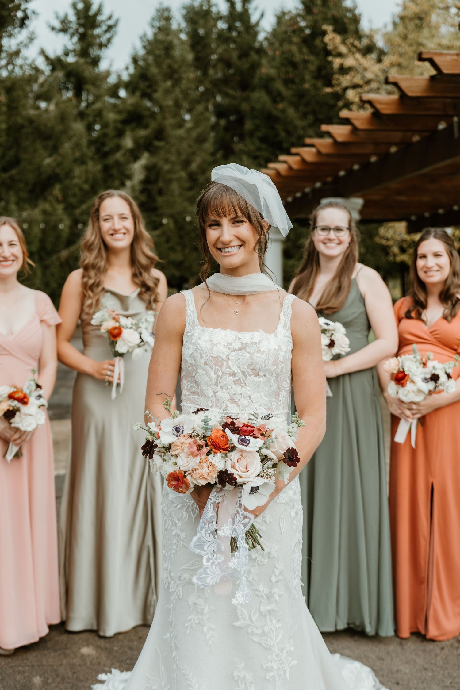 Smiling bride in lace gown holds colorful bouquet with bridesmaids in sage and coral dresses behind her outdoors.