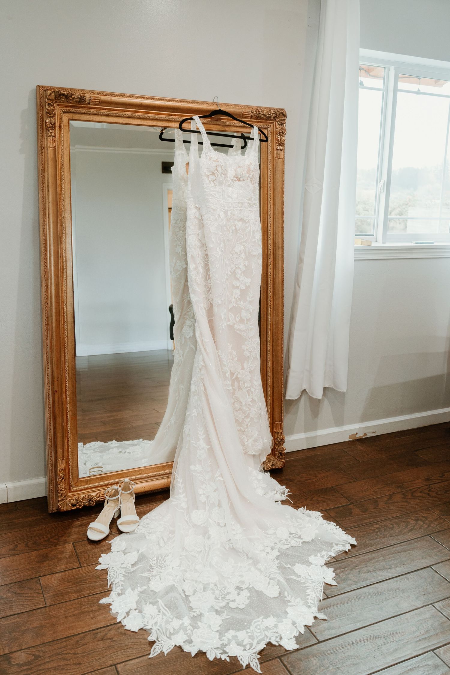 Elegant lace wedding dress with train hanging on gold ornate mirror with bridal heels on hardwood floor.