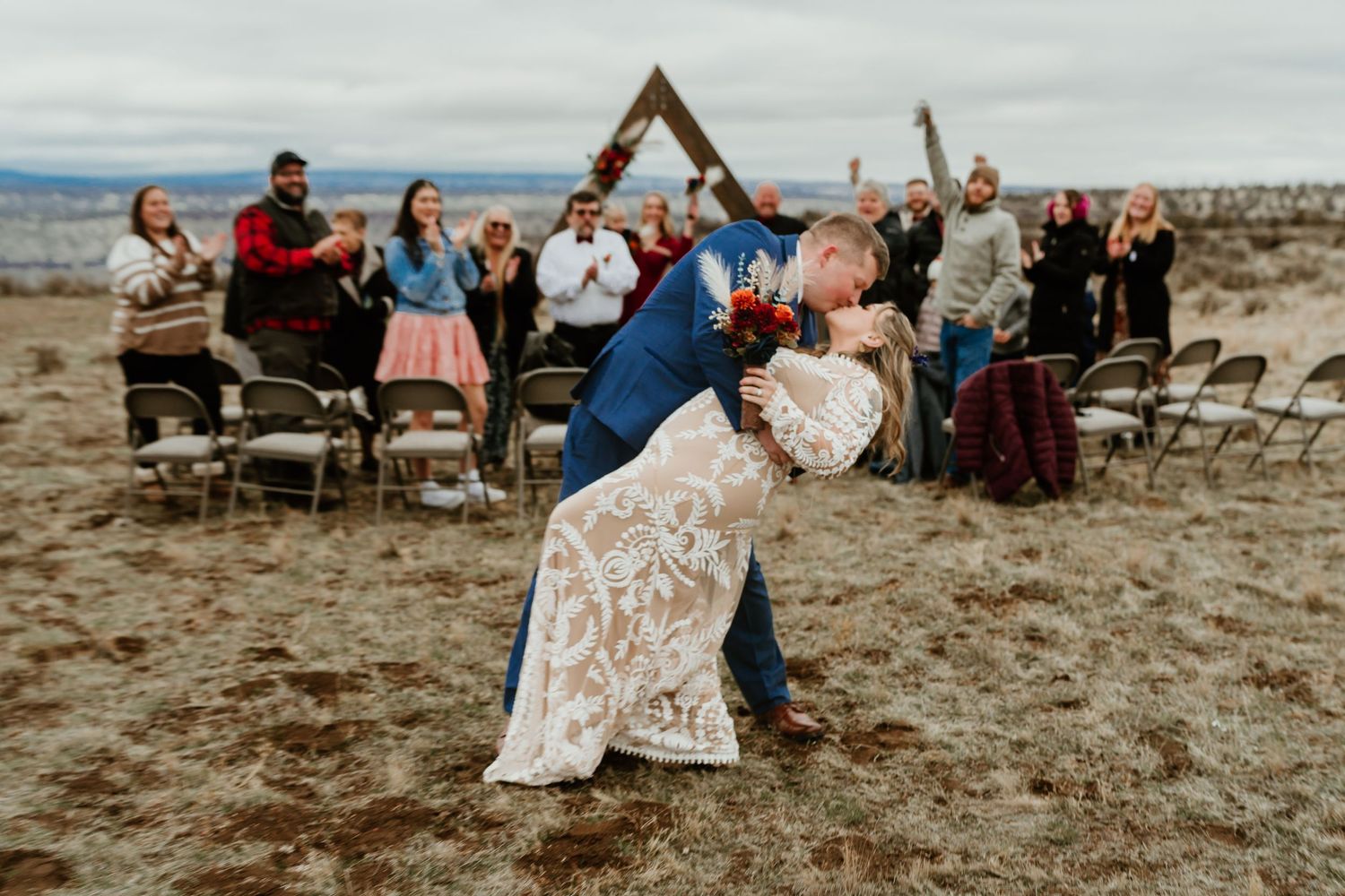 Groom dips bride in romantic first kiss at outdoor rustic wedding ceremony with cheering guests and triangular arch backdrop.