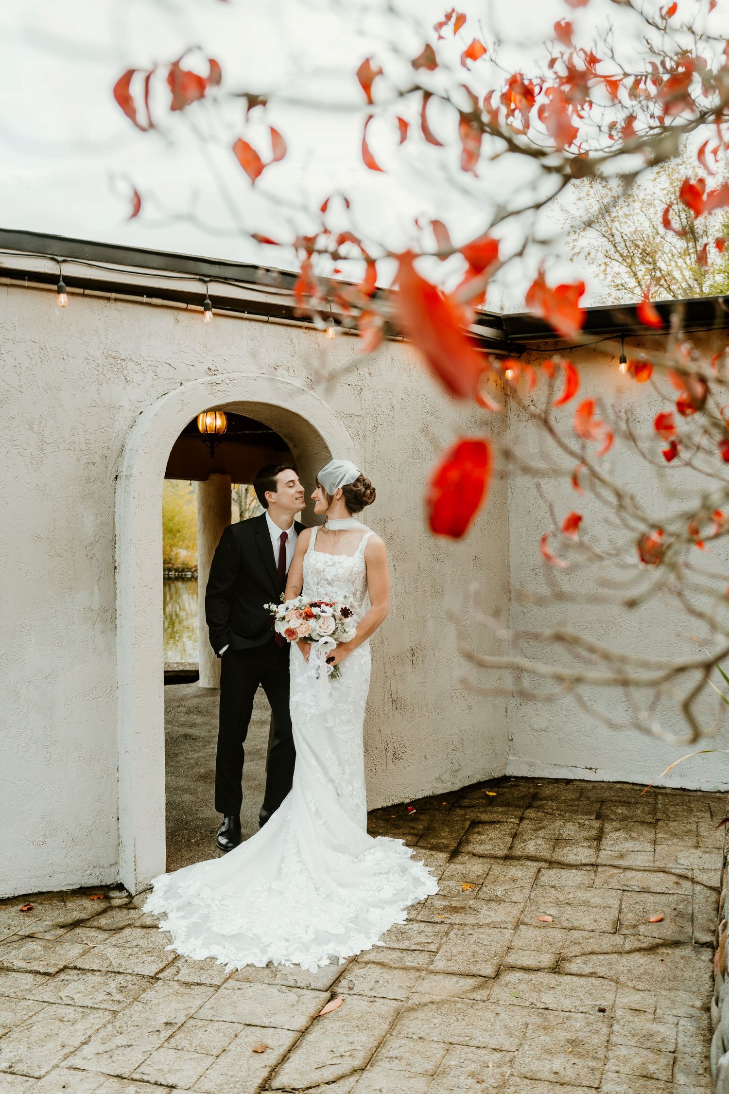 Bride and groom share a romantic moment under an arched doorway surrounded by vibrant red autumn leaves.