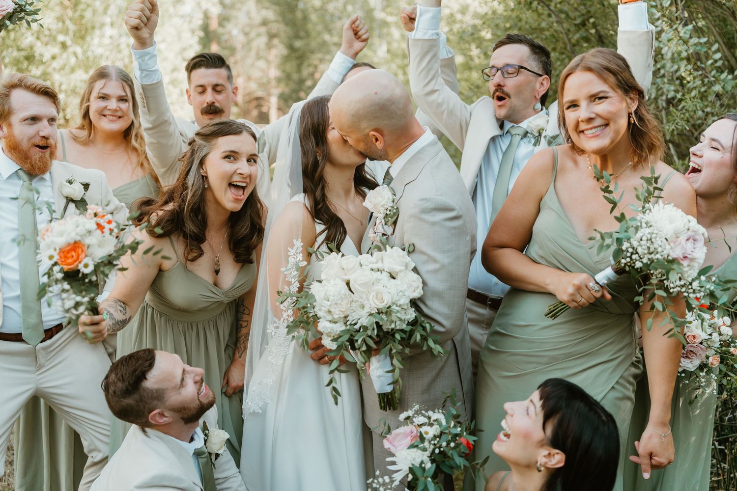 Joyful wedding party celebrates as bride and groom share a kiss, bridesmaids in sage green dresses holding white bouquets.
