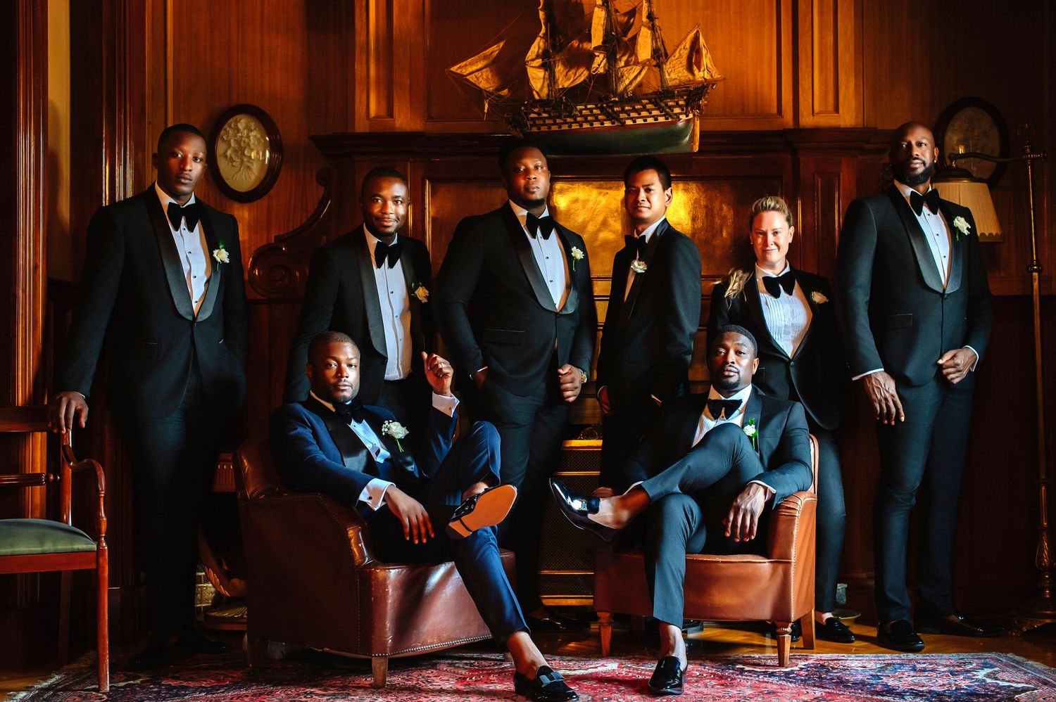 A formal group in black tuxedos poses together in an elegant wood-paneled room with leather chairs and a model ship display.