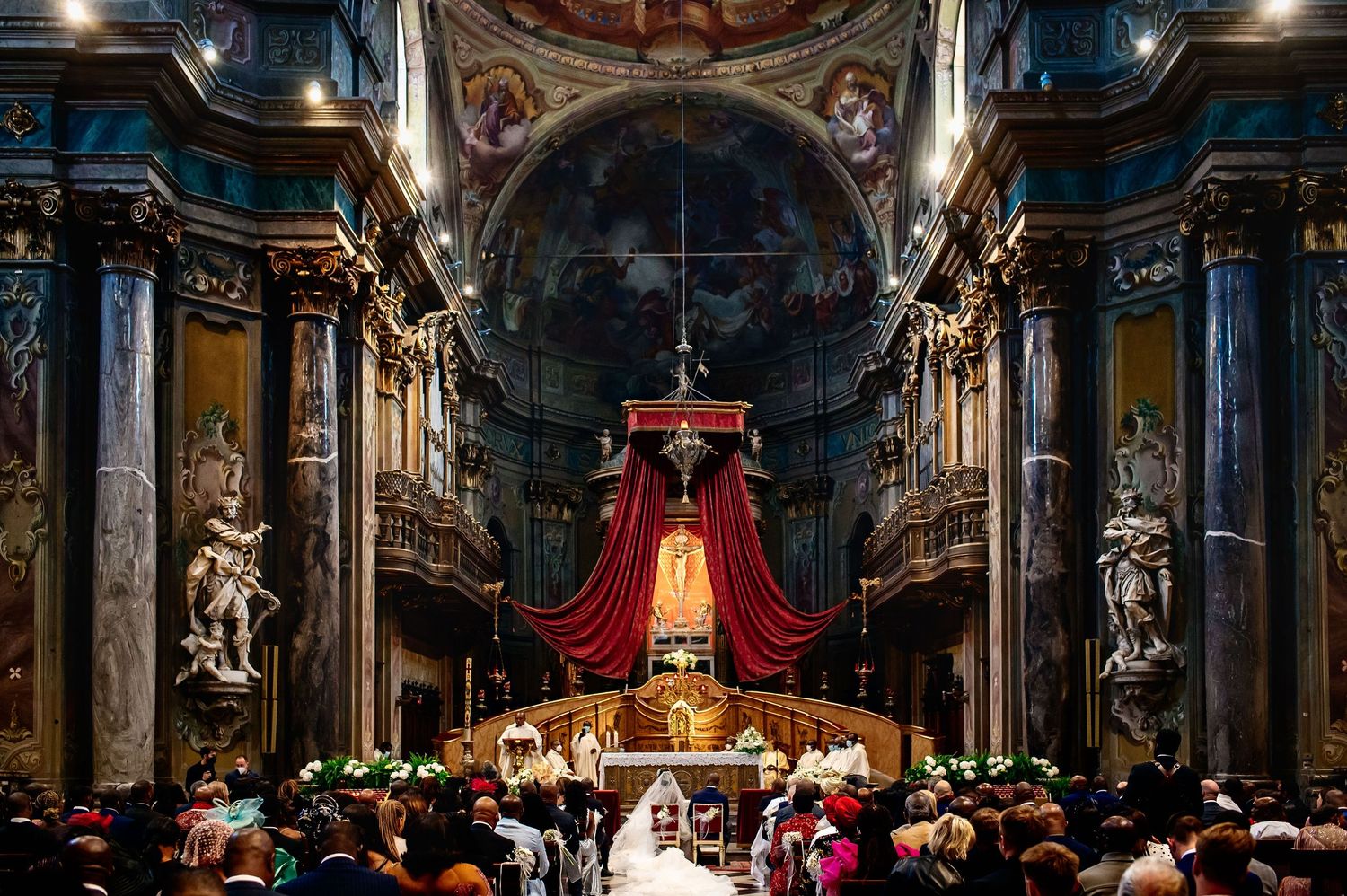 Ornate baroque church interior with red canopy and gathered crowd during a ceremony.