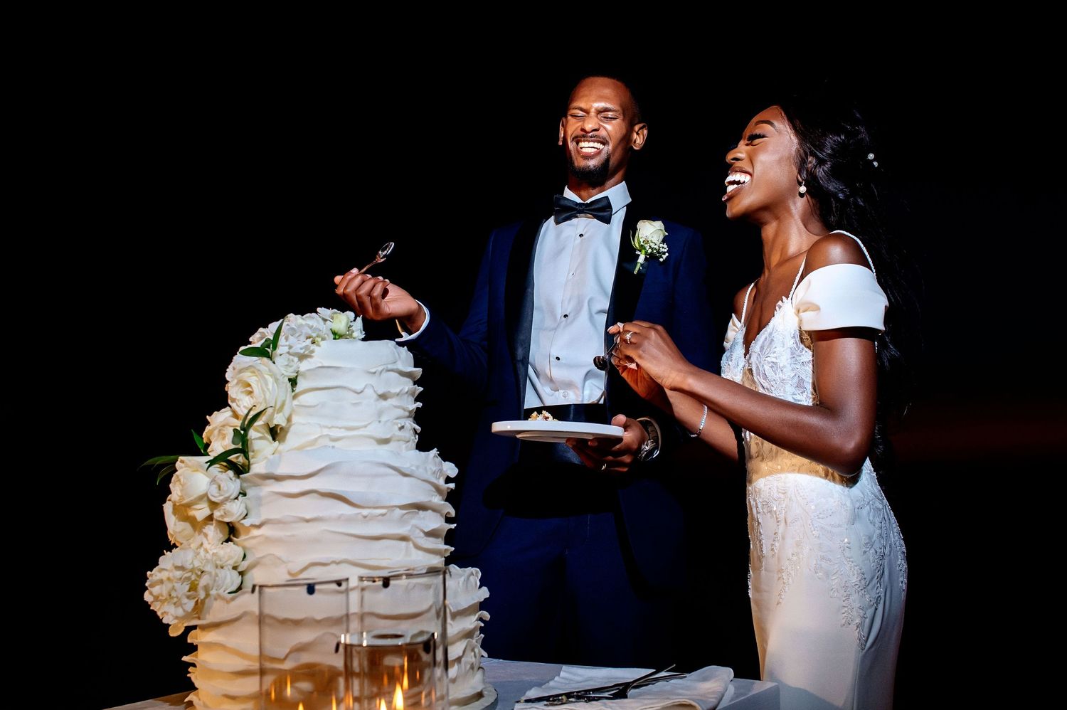 A couple shares a joyful moment while cutting their elegant white wedding cake decorated with delicate flowers.