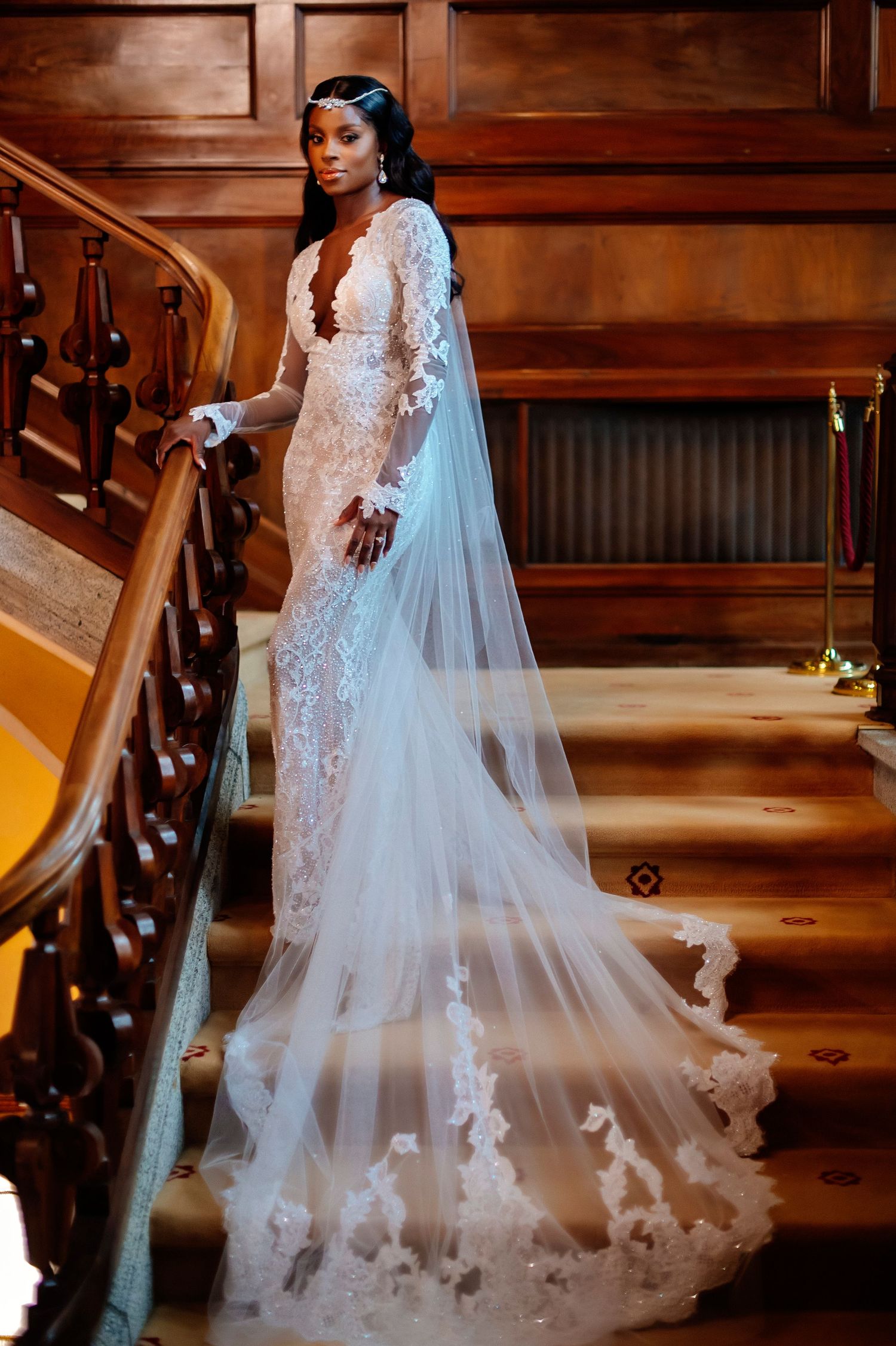 A stunning white lace wedding gown with a flowing cathedral veil is showcased on wooden stairs in warm lighting.