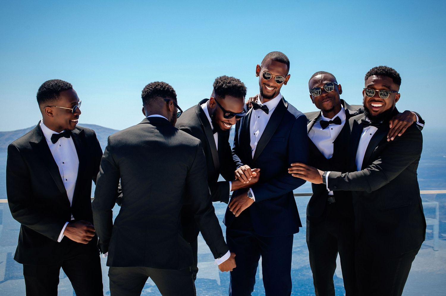 Groomsmen in black suits and bowties share a candid laughing moment together against a bright blue sky backdrop.