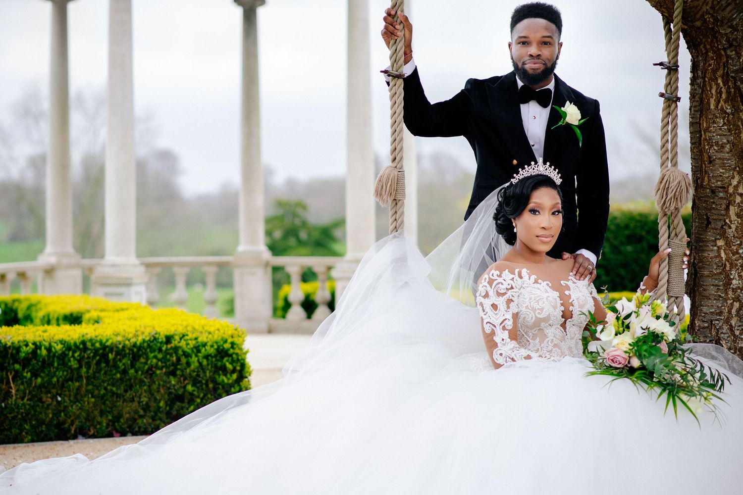 A couple poses elegantly in formal wedding attire near white columns and manicured gardens.