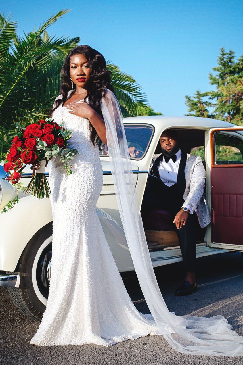 Bride in flowing white gown with red bouquet poses by vintage car.