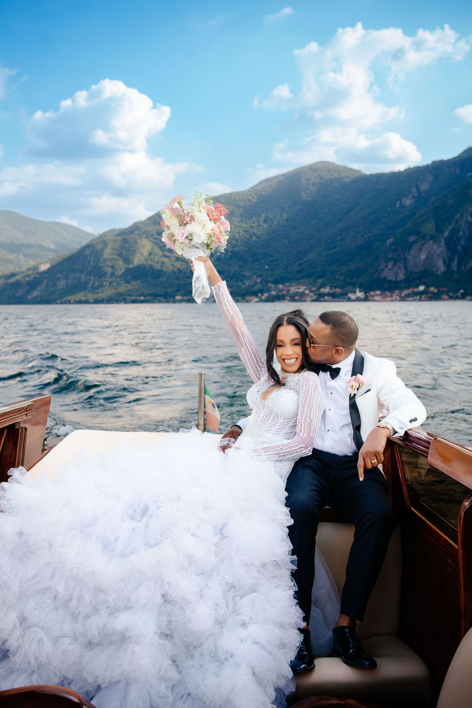 Wedding couple poses on a wooden boat with mountains and lake in the background.