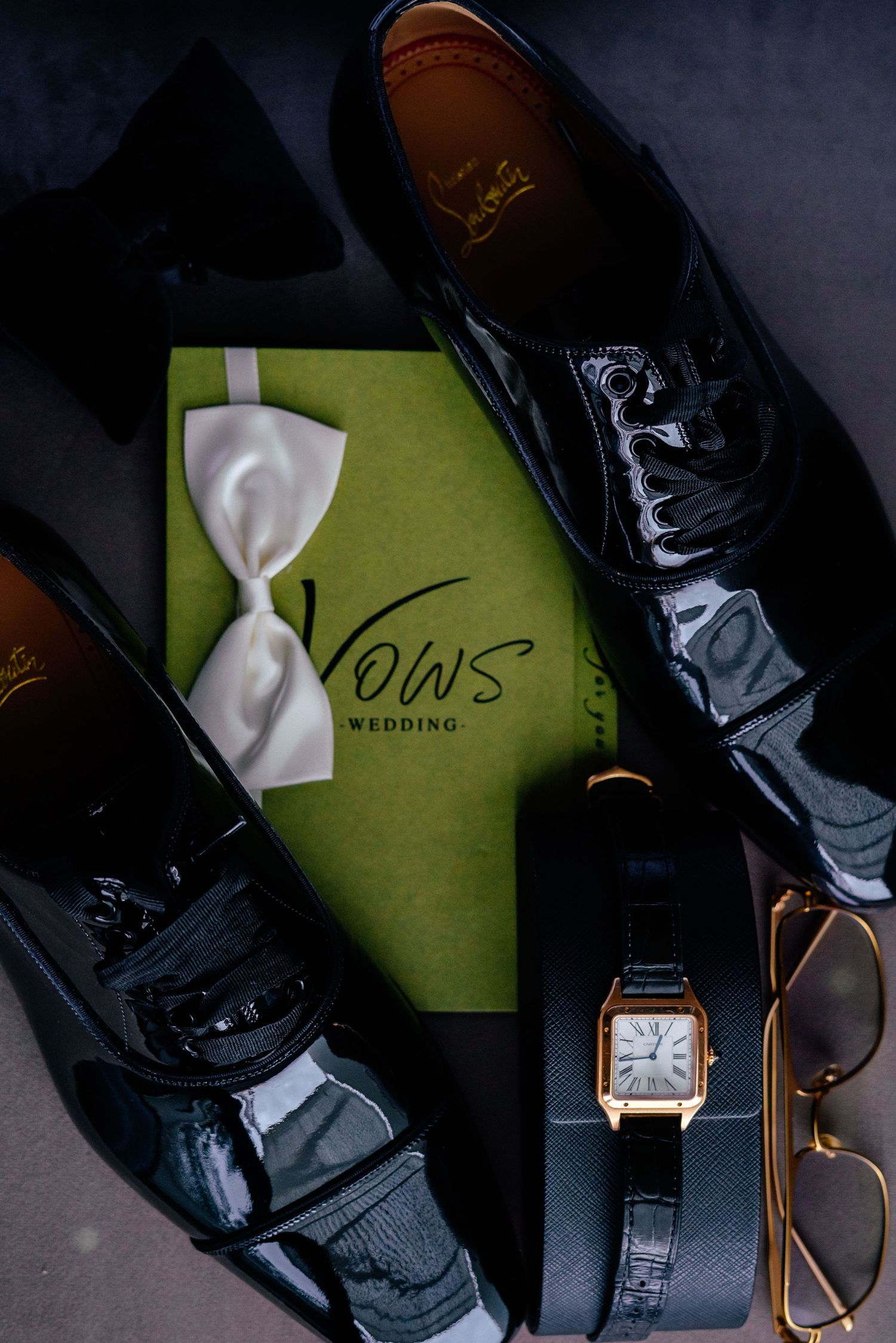 Wedding accessories including dress shoes, watch, and vows book arranged on dark surface.