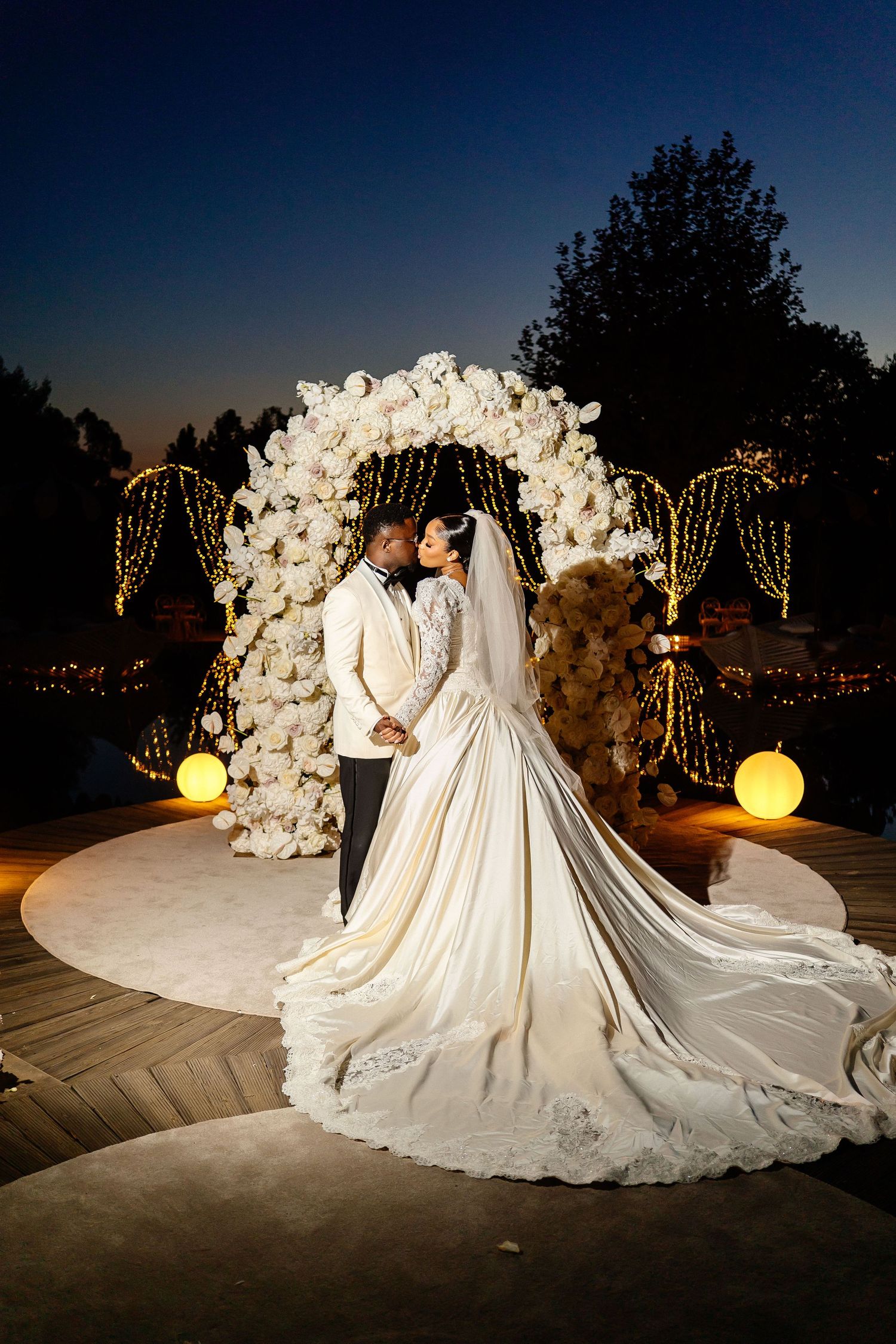 A romantic nighttime wedding scene under a floral arch with glowing lights and white roses.