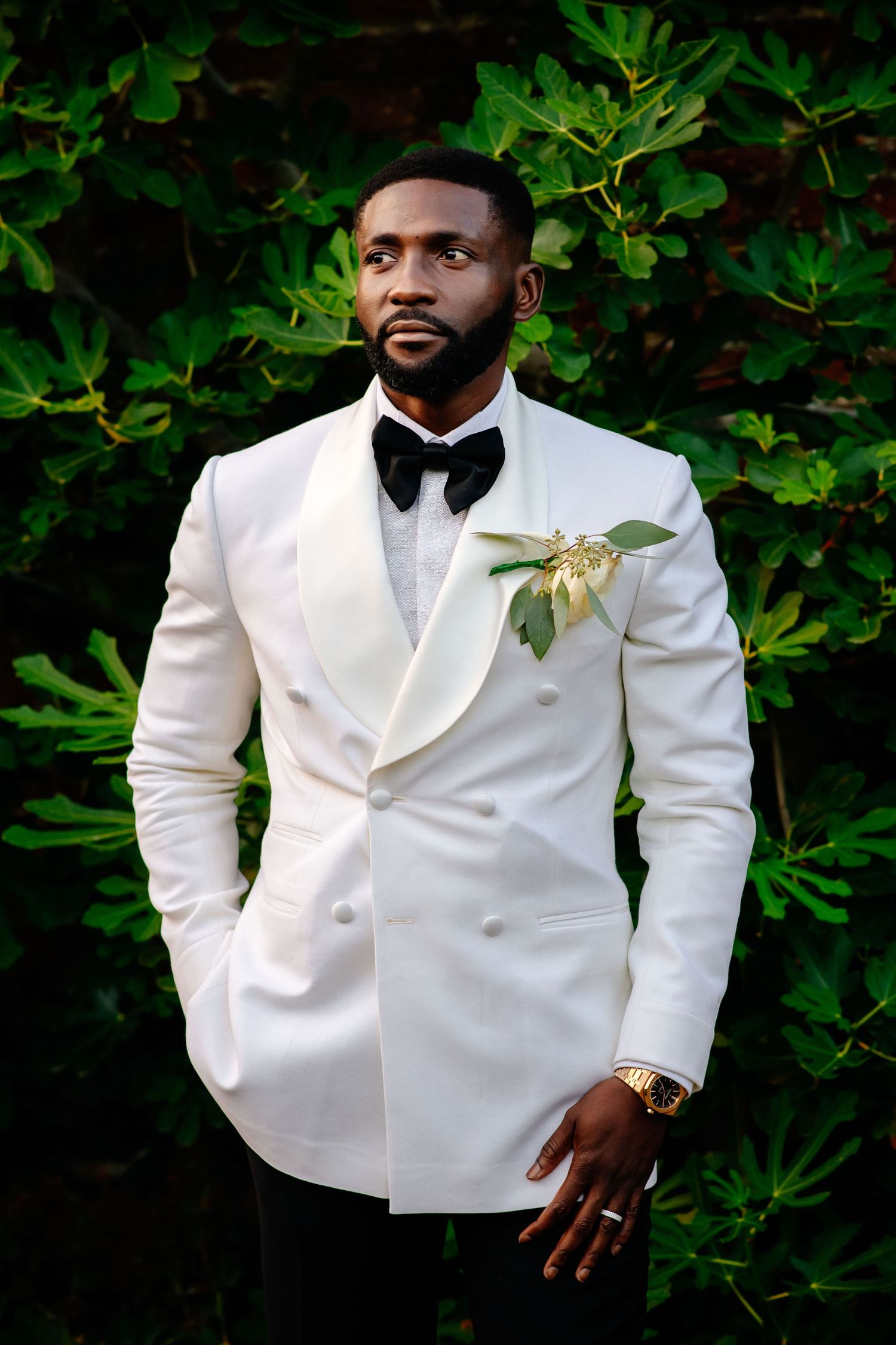 Elegant white tuxedo jacket with black bow tie and floral boutonniere against lush green foliage background.