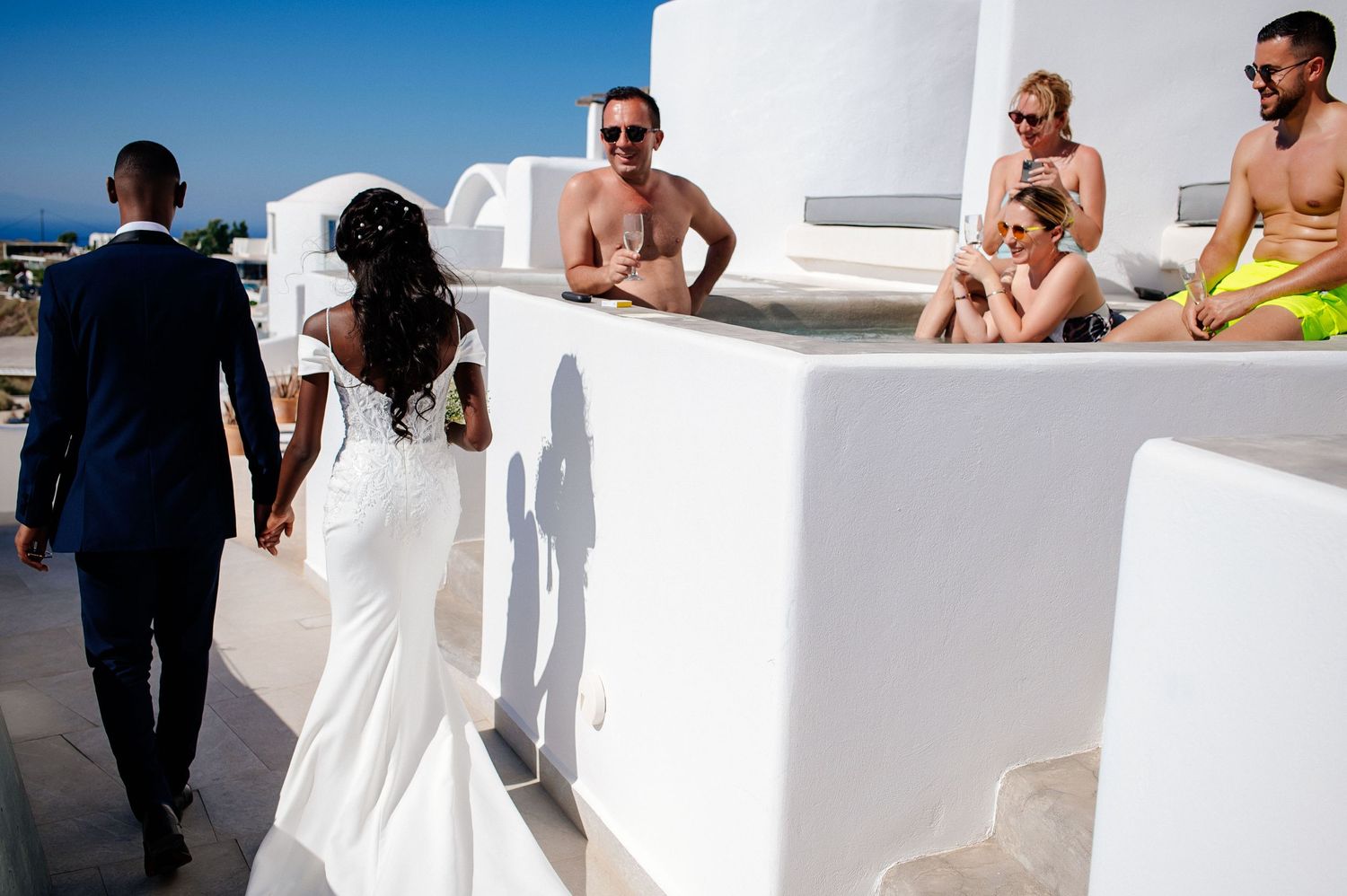A group of people in formal wedding attire walking along a white stucco wall on a sunny Mediterranean day.