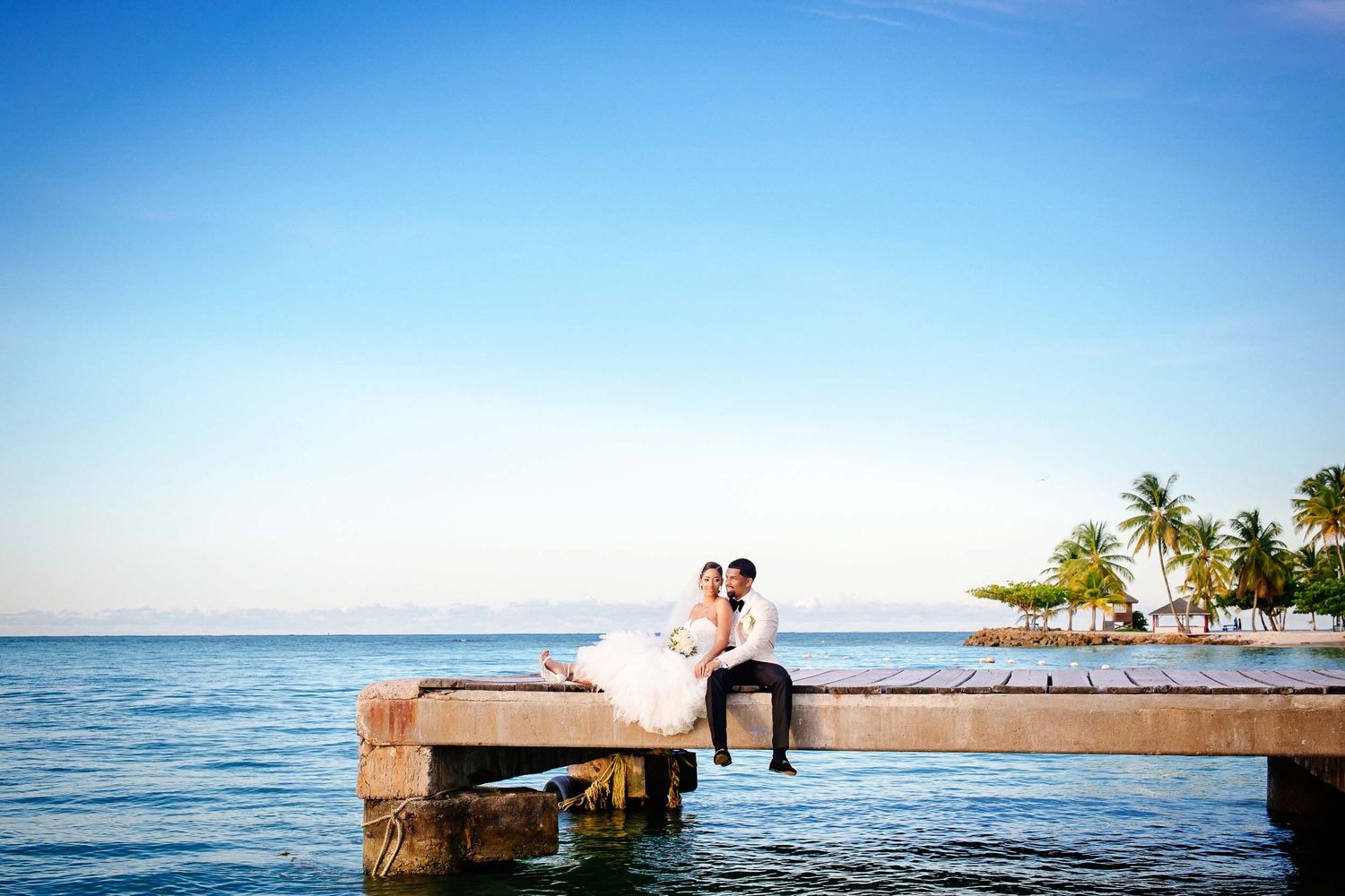 Couple sitting on wooden pier by ocean at sunset with palm trees in background.