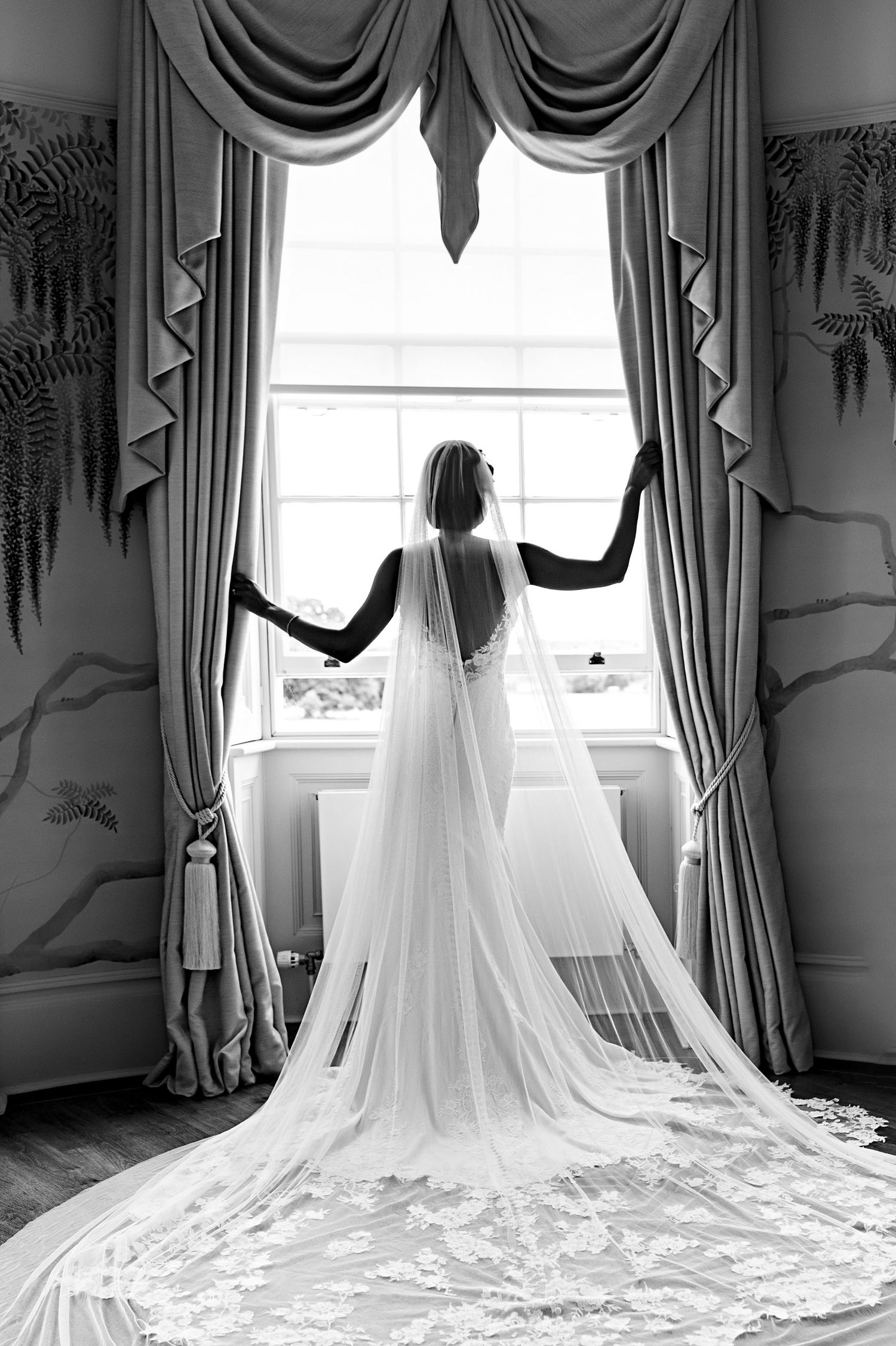 A silhouette of a bride in a flowing cathedral veil stands dramatically by an ornate window.
