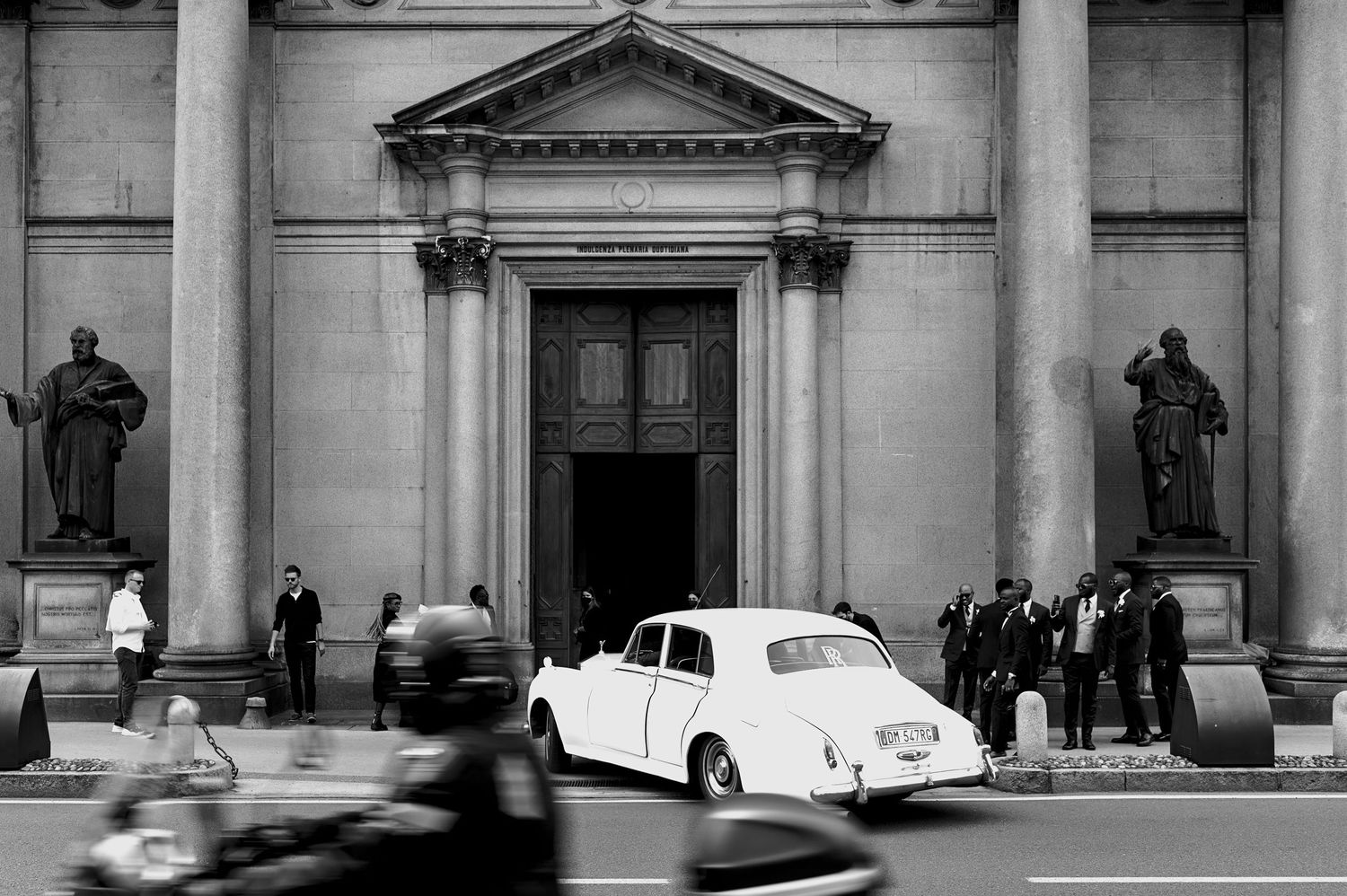 Vintage black and white photo of classic car parked outside grand building with columns.