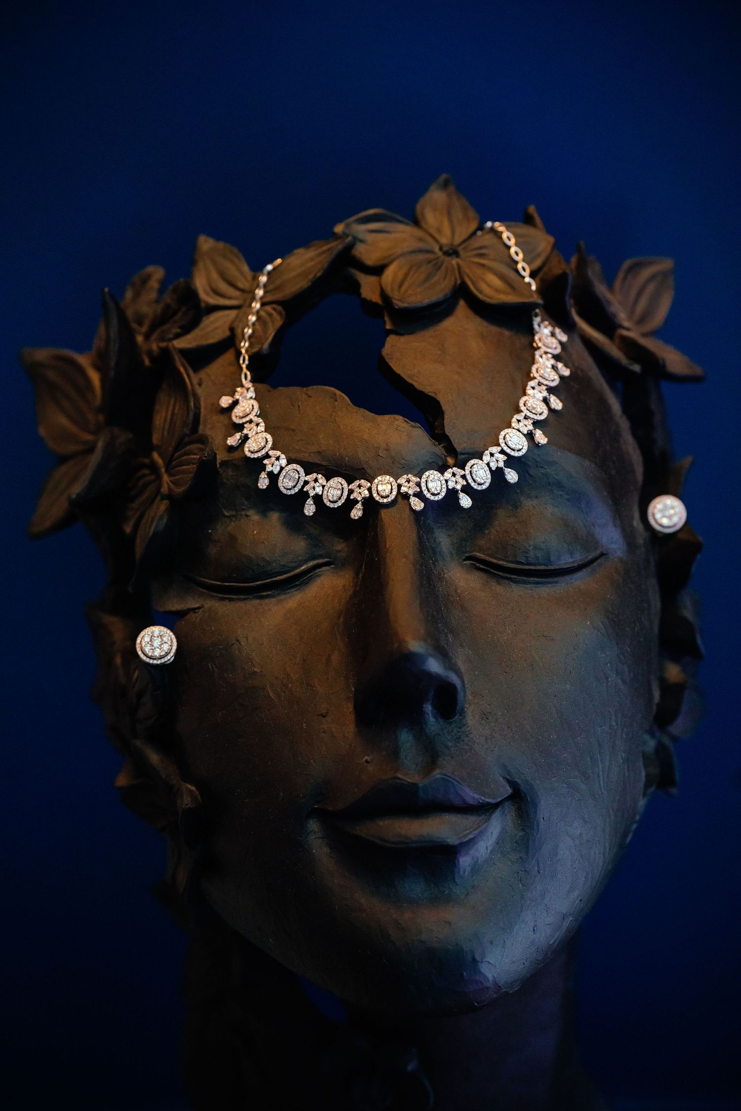 Artistic close-up of decorative statue wearing delicate crystal jewelry against dark blue background.