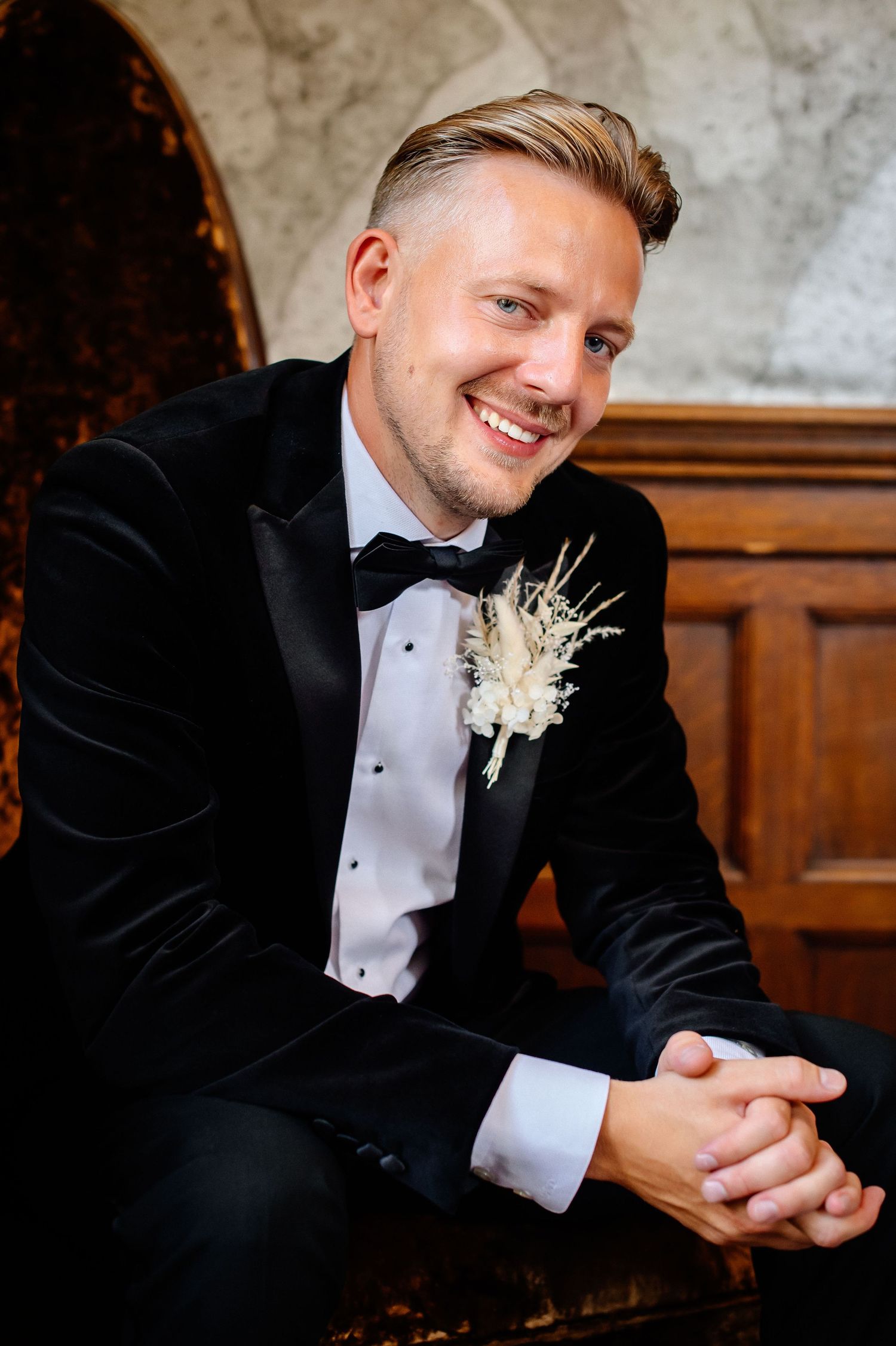 Groom in black tuxedo and white boutonniere smiles during formal wedding portraits.