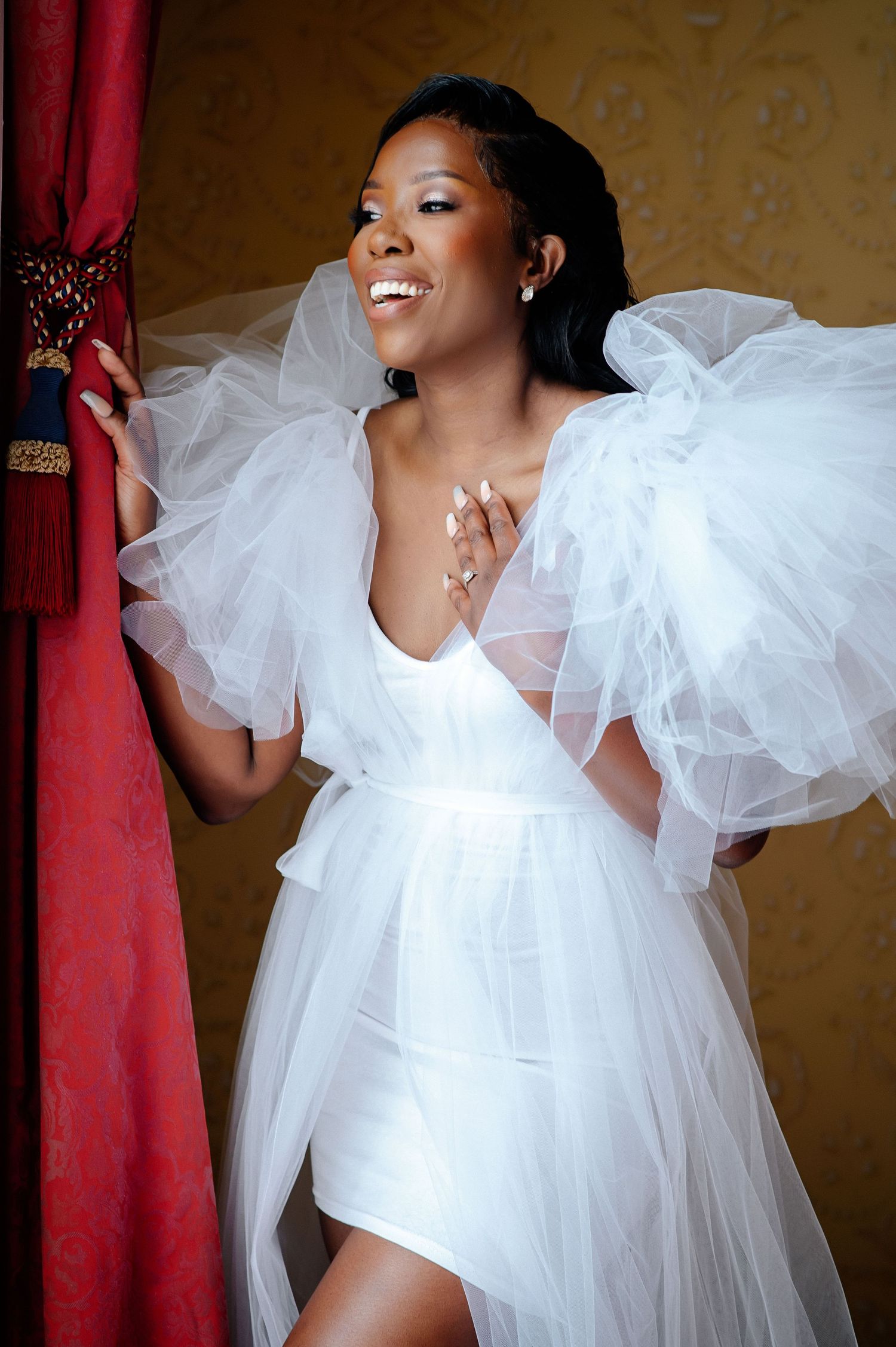 Bride in flowing white tulle dress poses against red wall.