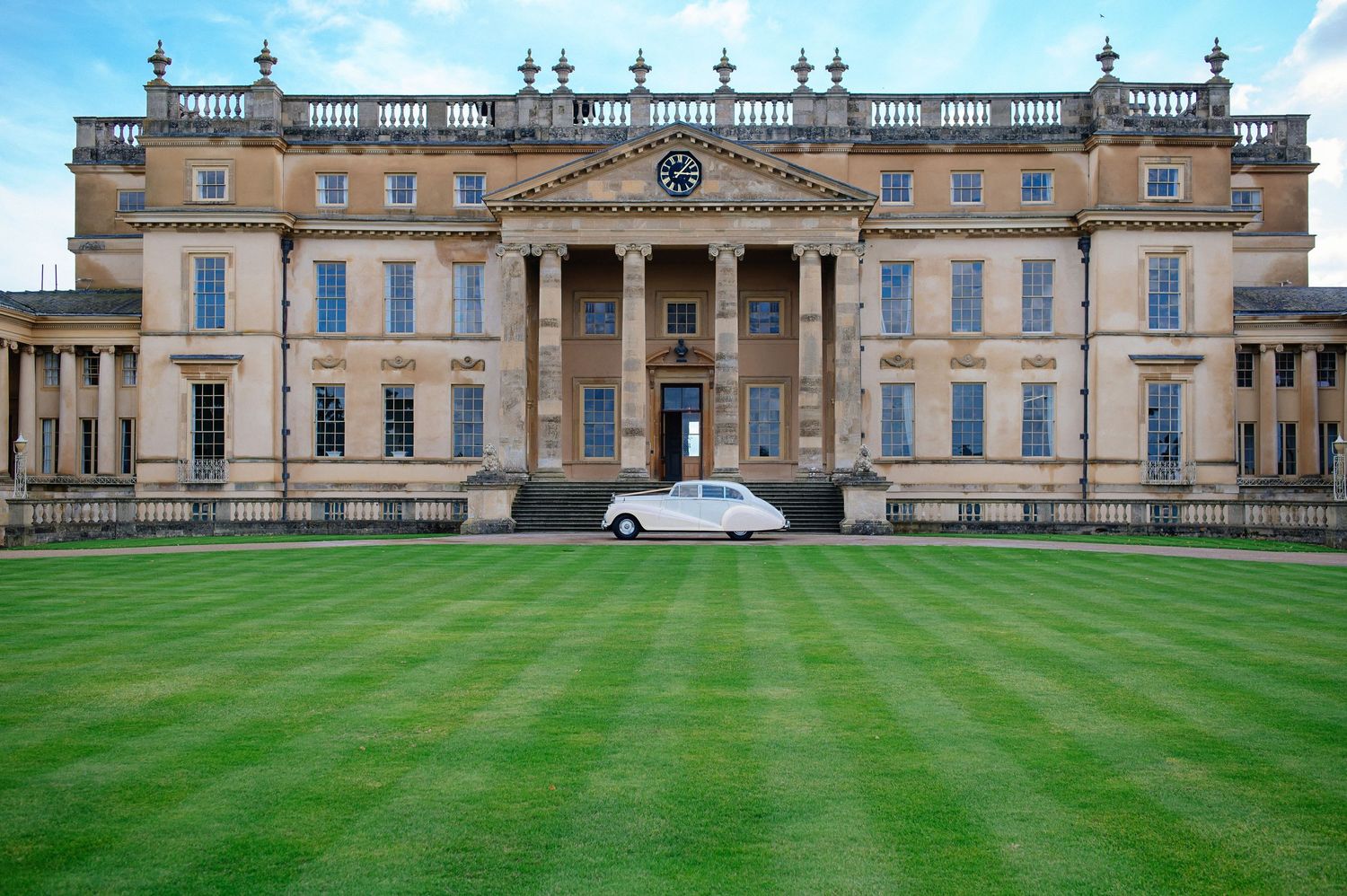 Panoramic view of a grand historic mansion with ornate architecture and manicured green lawns stretching across the facade.