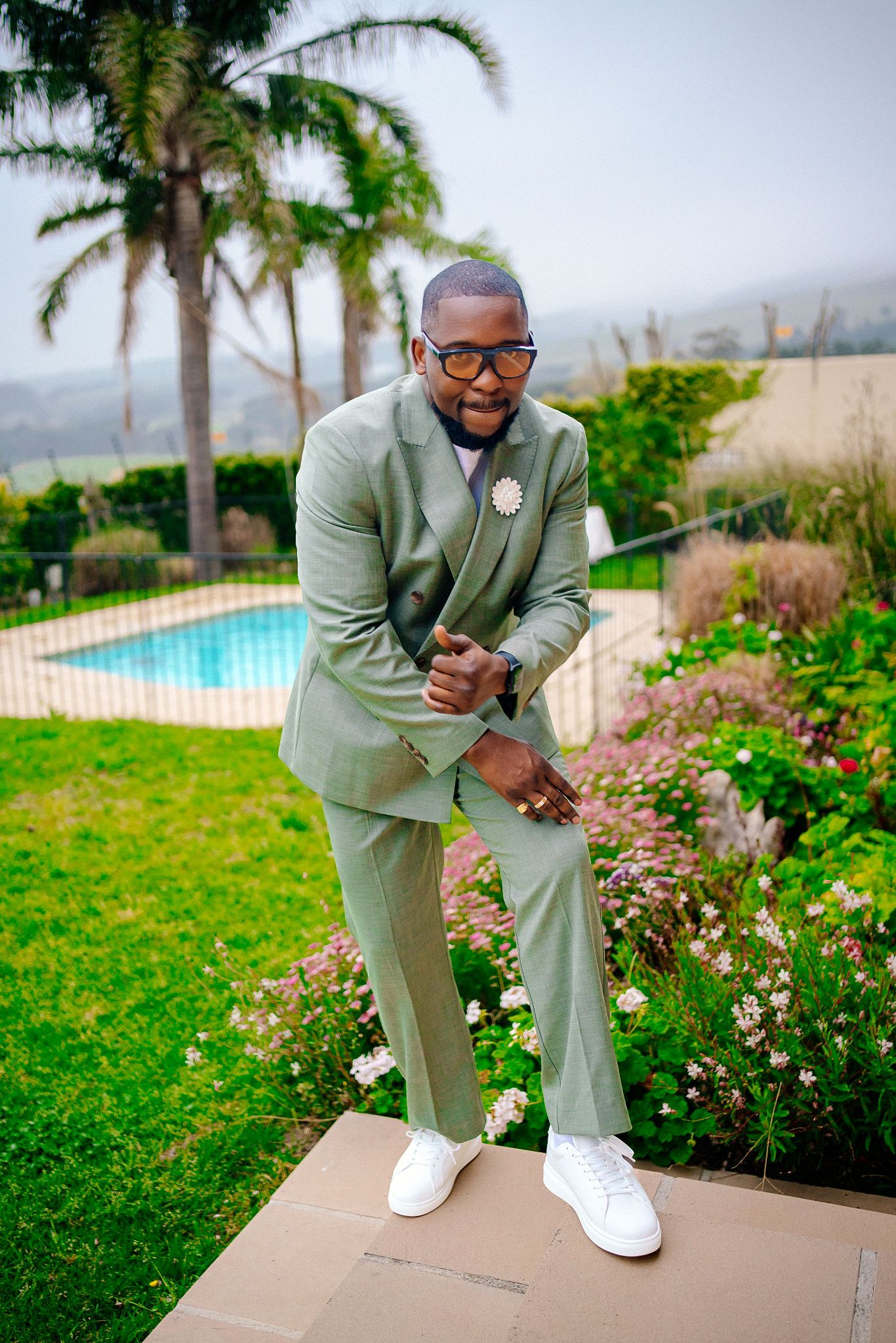A person in a mint green suit sits on a ledge near a pool surrounded by palm trees and tropical landscaping.