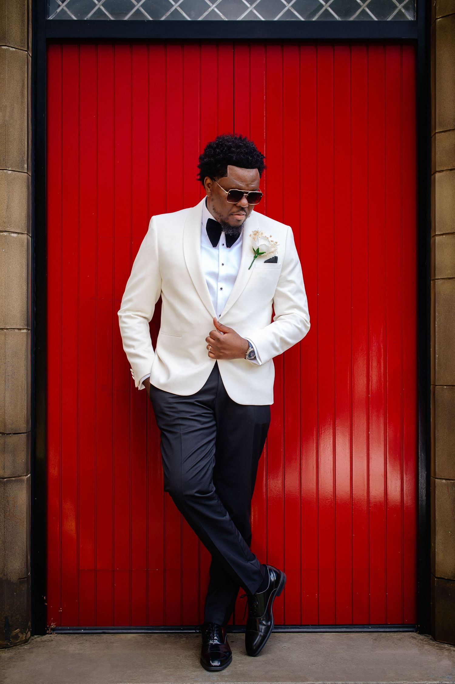 Stylish groom in white tuxedo jacket poses against vibrant red doors for wedding portraits.