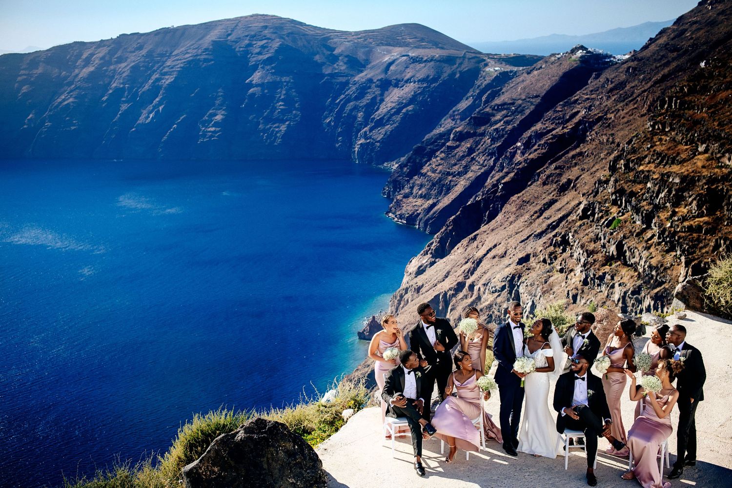 A wedding ceremony takes place on a cliffside overlooking dramatic blue Mediterranean waters.