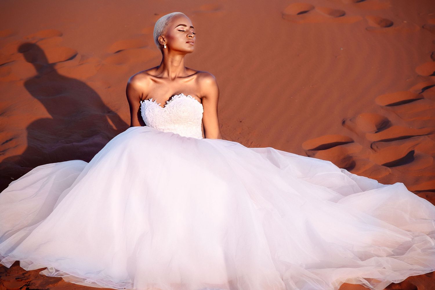 Bride in white wedding gown poses dramatically against red desert sand dunes.