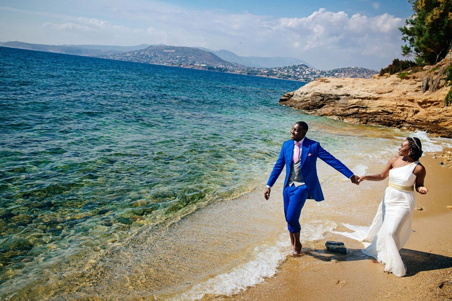 A couple in wedding attire walks hand in hand along a beautiful Mediterranean beach with crystal clear turquoise waters.
