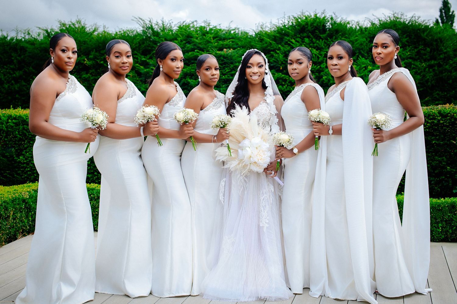 A group of bridesmaids in white dresses pose together outdoors in a garden setting.