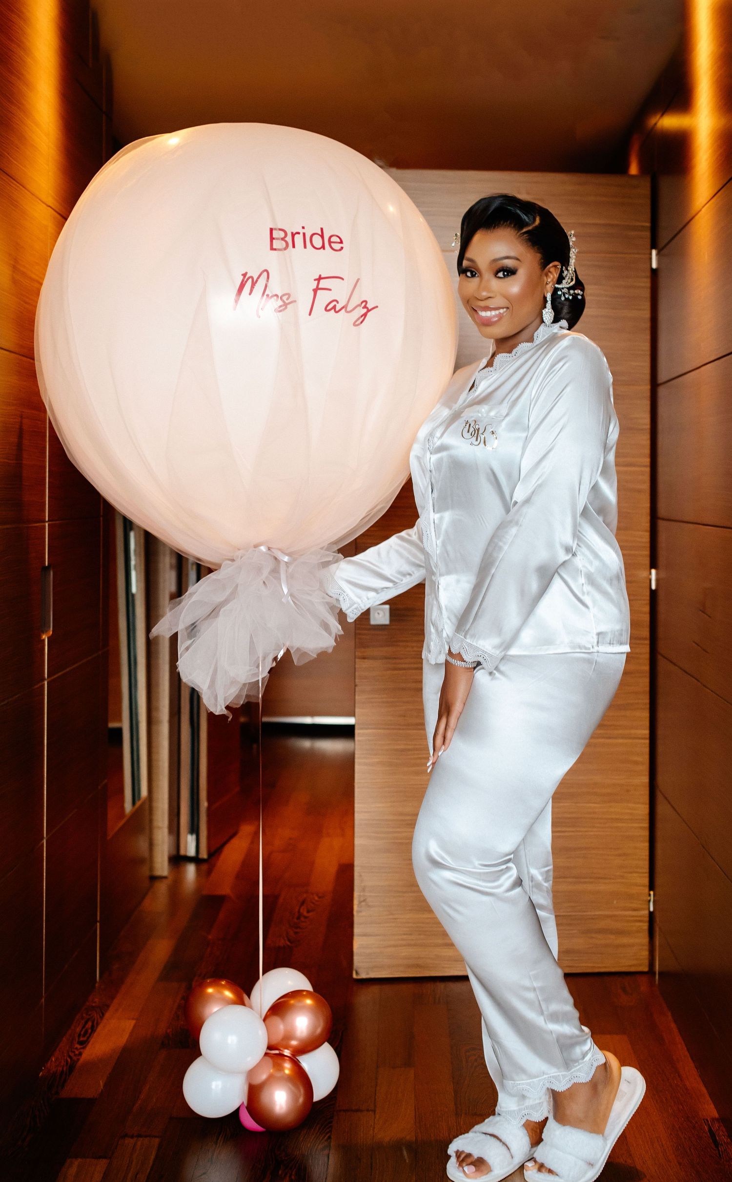 Bride in white silk pajamas poses with custom balloons during wedding preparation.