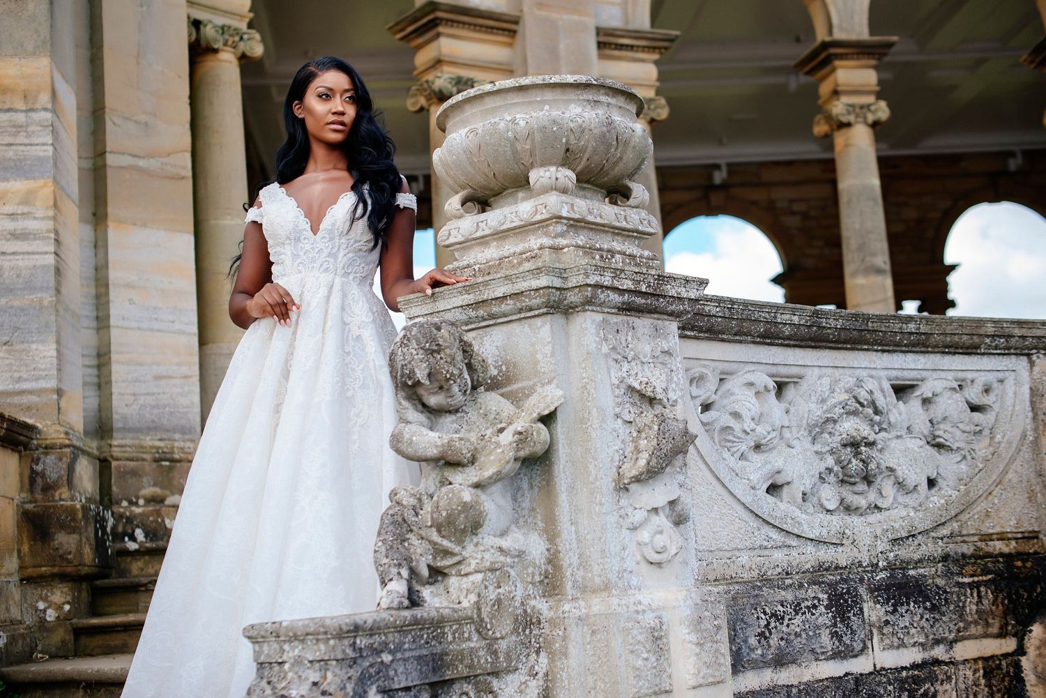 A bride in a flowing white ballgown poses on ornate stone steps of a classical building with decorative architecture.