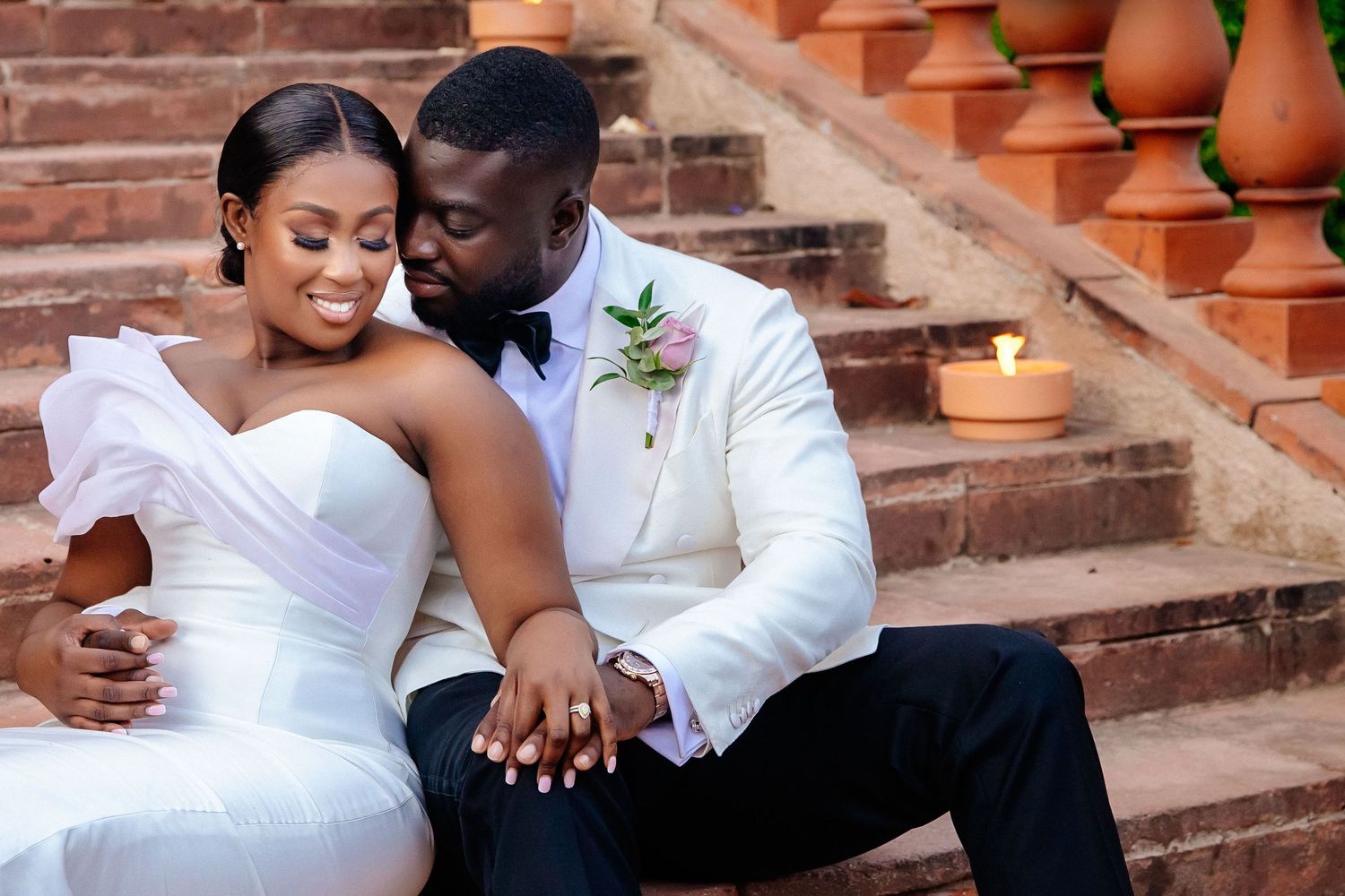 A couple in white outfits sits intimately on brick steps decorated with terracotta pots.