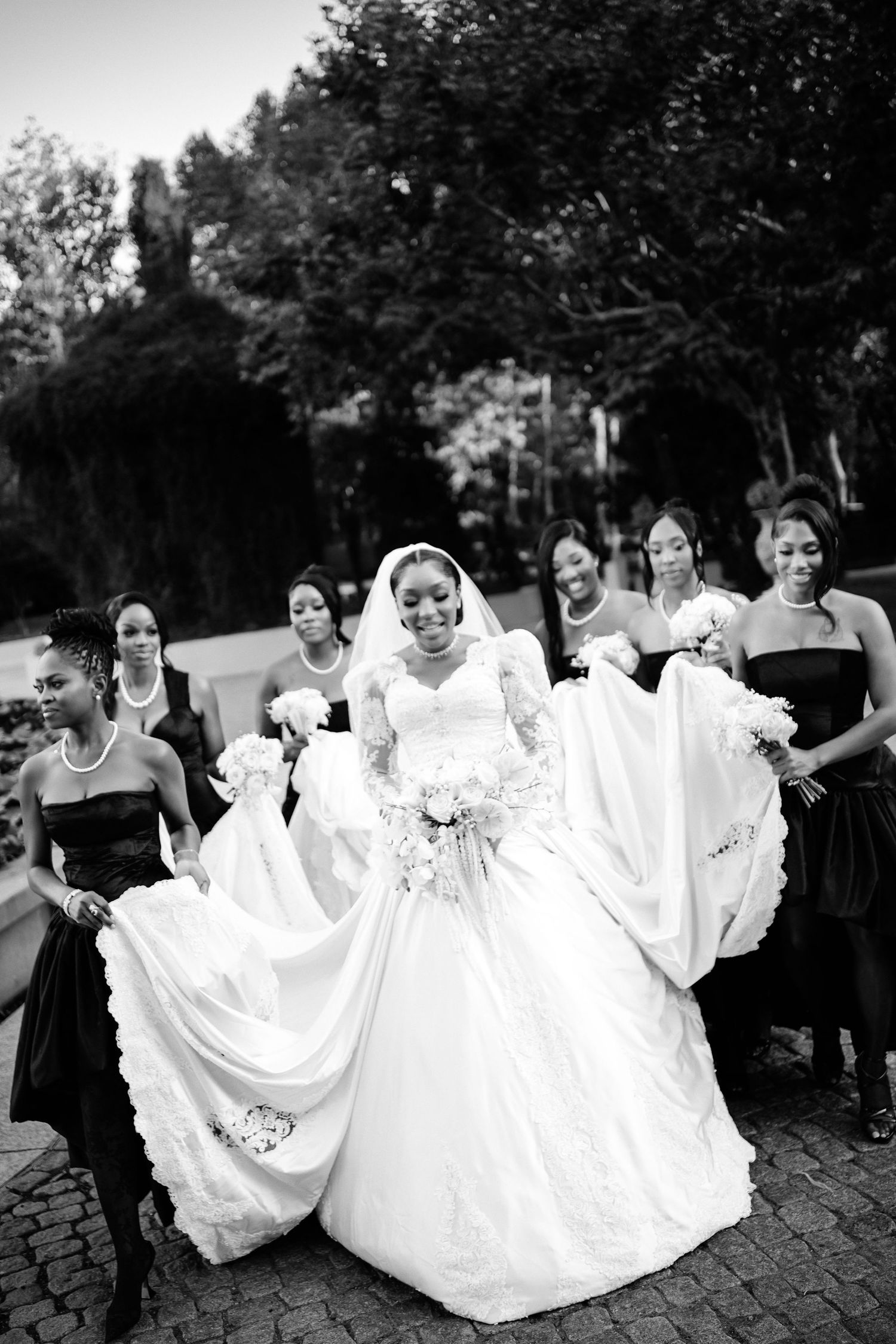 Joyful bridesmaids in black dresses playfully lifting white gowns in outdoor setting.