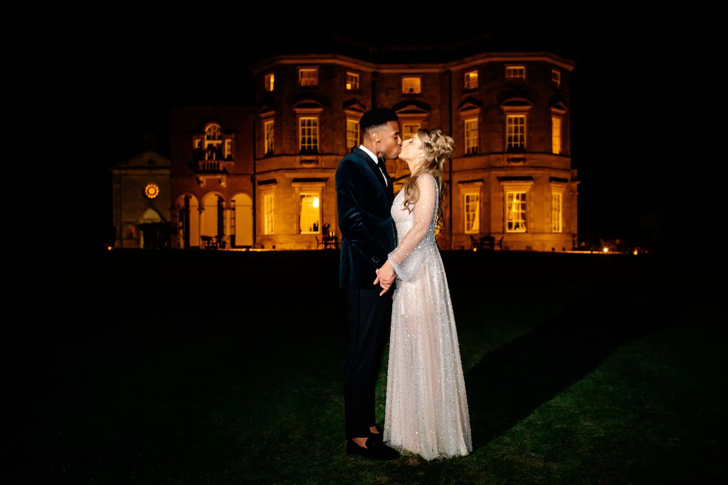 A romantic nighttime wedding portrait in front of an illuminated historic mansion.