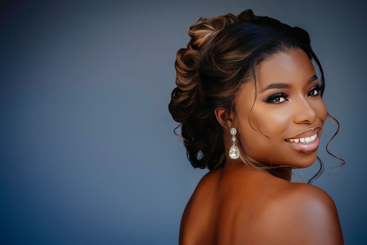 Portrait of a smiling woman with an elegant updo hairstyle and pearl drop earrings against a blue background.